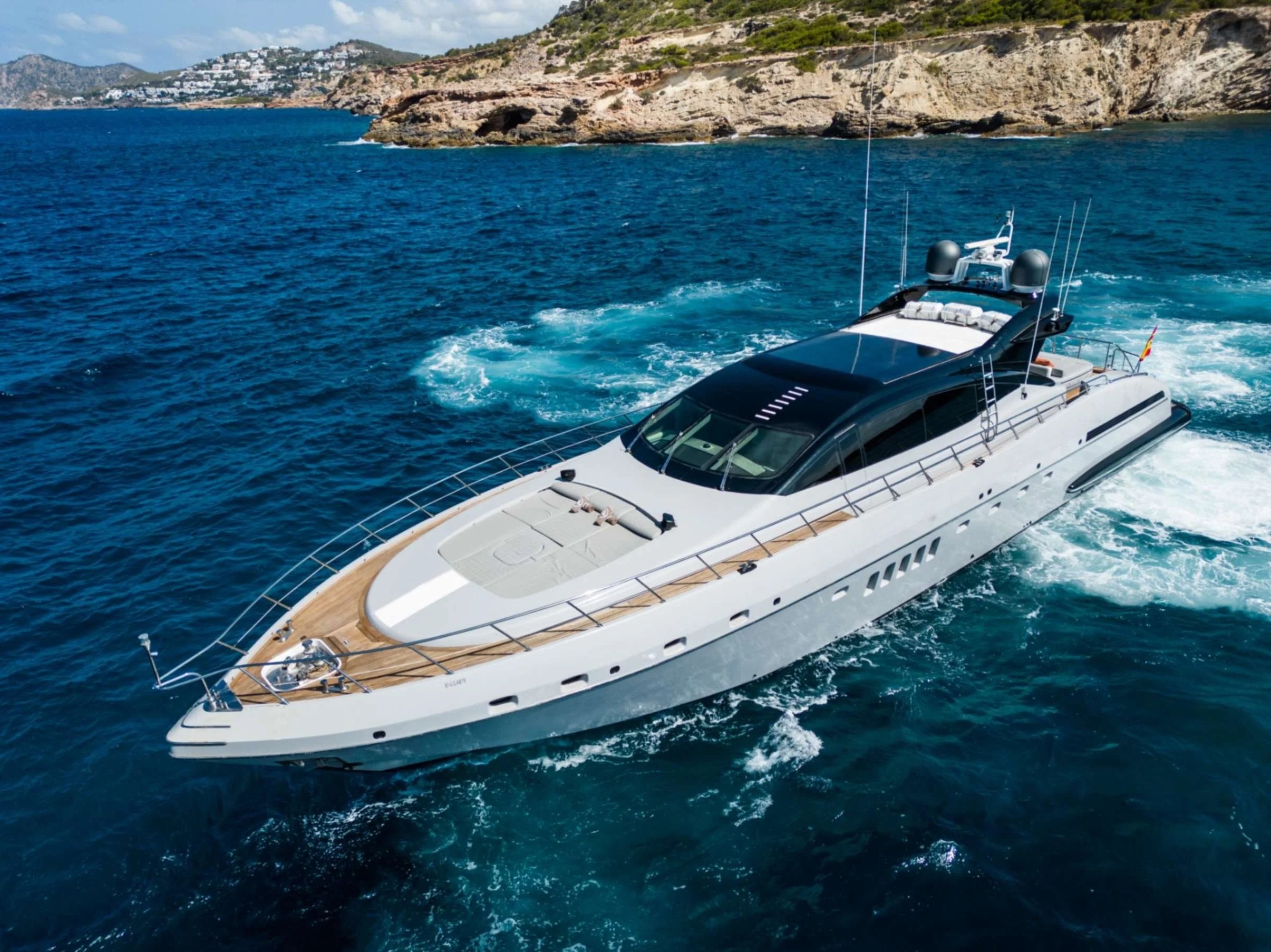 Luxury yacht sailing in blue ocean near rocky coastline with buildings on the hillside.