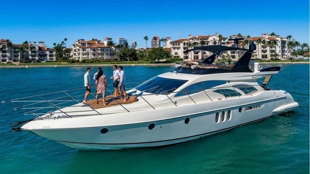 A luxury yacht with several people on the deck, cruising along a waterway with residential buildings and palm trees in the background on a clear, sunny day.