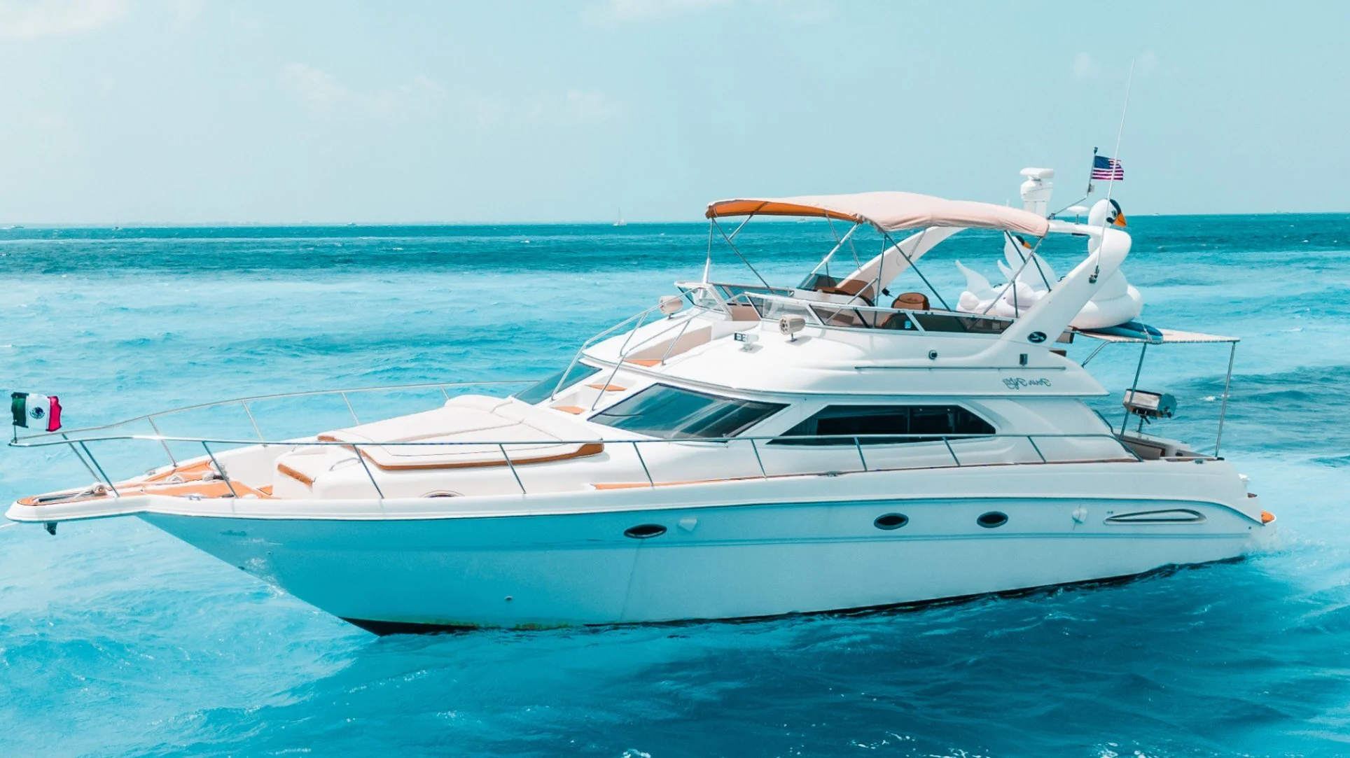A white yacht with a beige canopy sailing on blue ocean waters with an American flag on top and a swan inflatable decoration at the back.