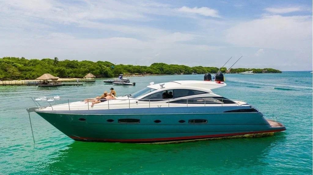 A large luxury yacht floating on clear turquoise water near a wooded shoreline with small huts and boats in the background, under a partly cloudy sky.