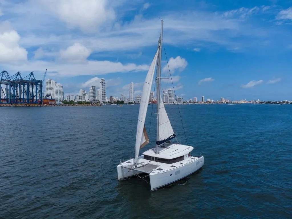 A white sailboat on calm water with a city skyline and blue sky in the background.