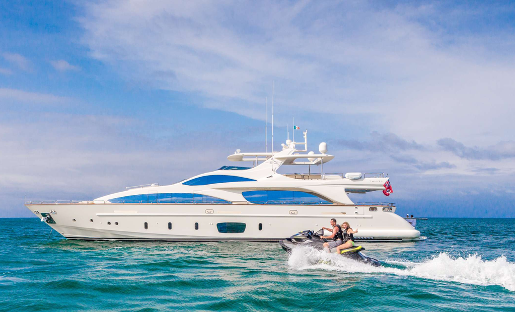 A large white yacht with blue accents on the water, with two people riding a Jet Ski nearby under a partly cloudy sky.