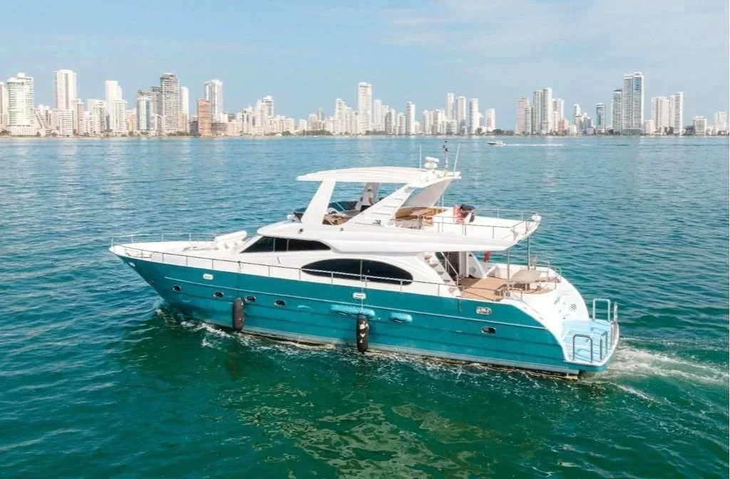 A white and turquoise yacht sailing in calm blue water with a city skyline of high-rise buildings in the background.