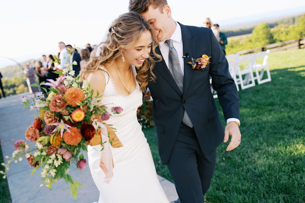 Hilltop ceremony with bride and groom walking outdoors during their wedding ceremony, smiling and leaning towards each other, with guests and chairs in the background.
