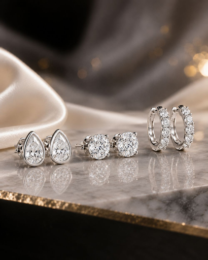 White gold diamond stud, pear, and huggie earrings displayed on a luxury marble surface with elegant lighting.