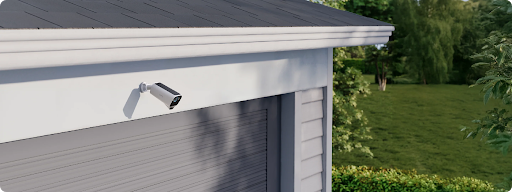 Outdoor security camera mounted under a garage eave overlooking a sunny green backyard