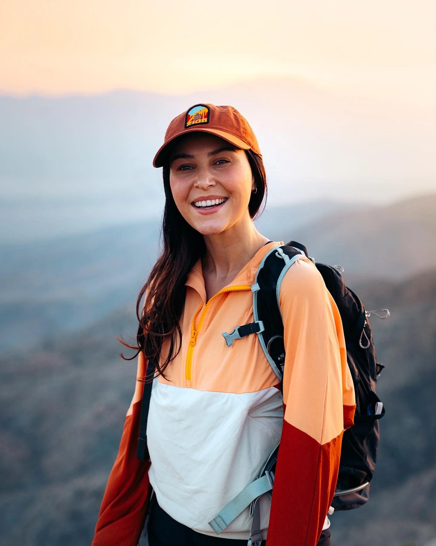 Young woman hiking outdoors at sunset, wearing a tan cap, orange jacket, and carrying a backpack.