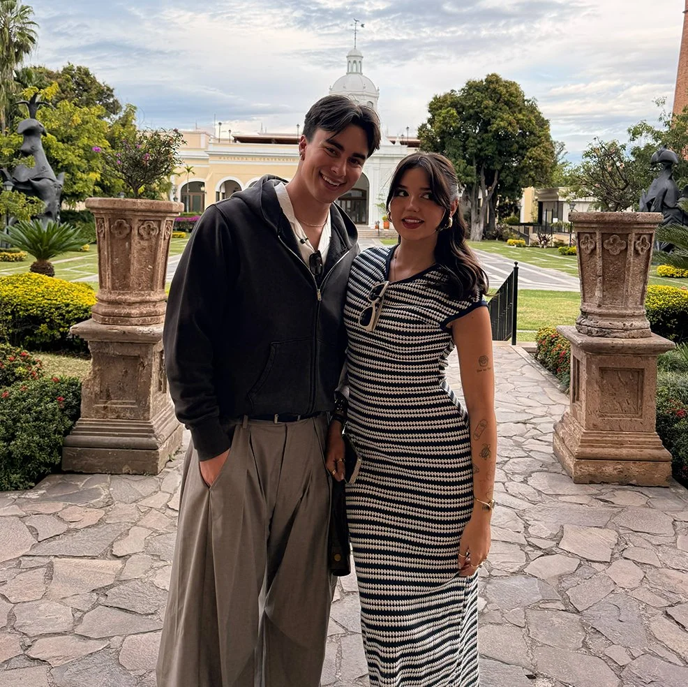 A young man and woman posing together outdoors on a stone pathway with a garden and historic building in the background.