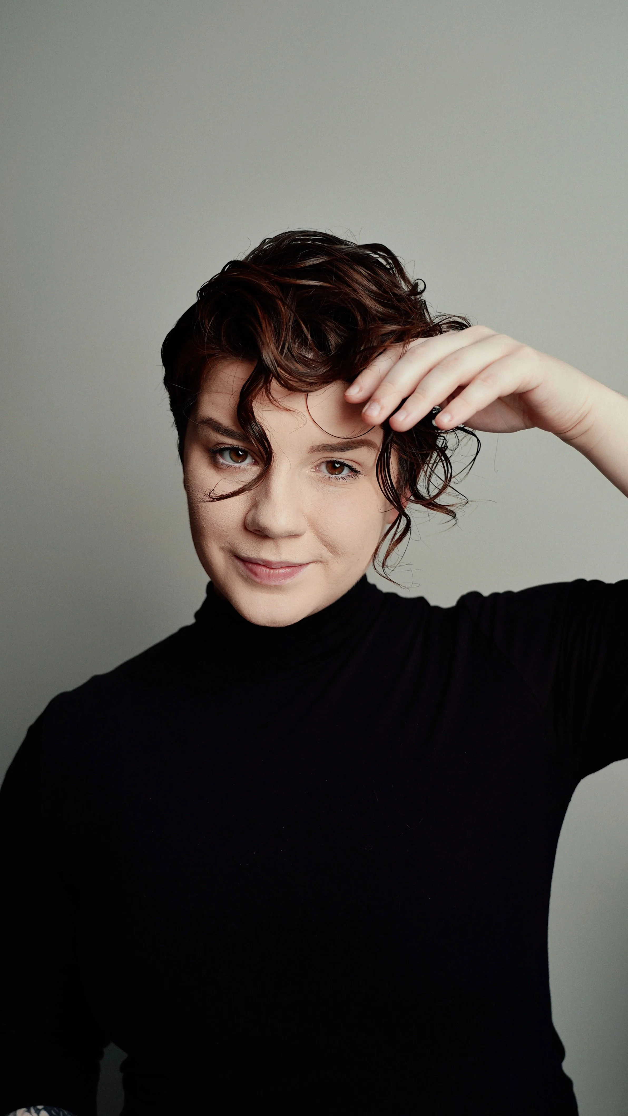 A woman with short, curly, dark red hair adjusting her hair with her right hand, wearing a black turtleneck, against a plain light gray background.