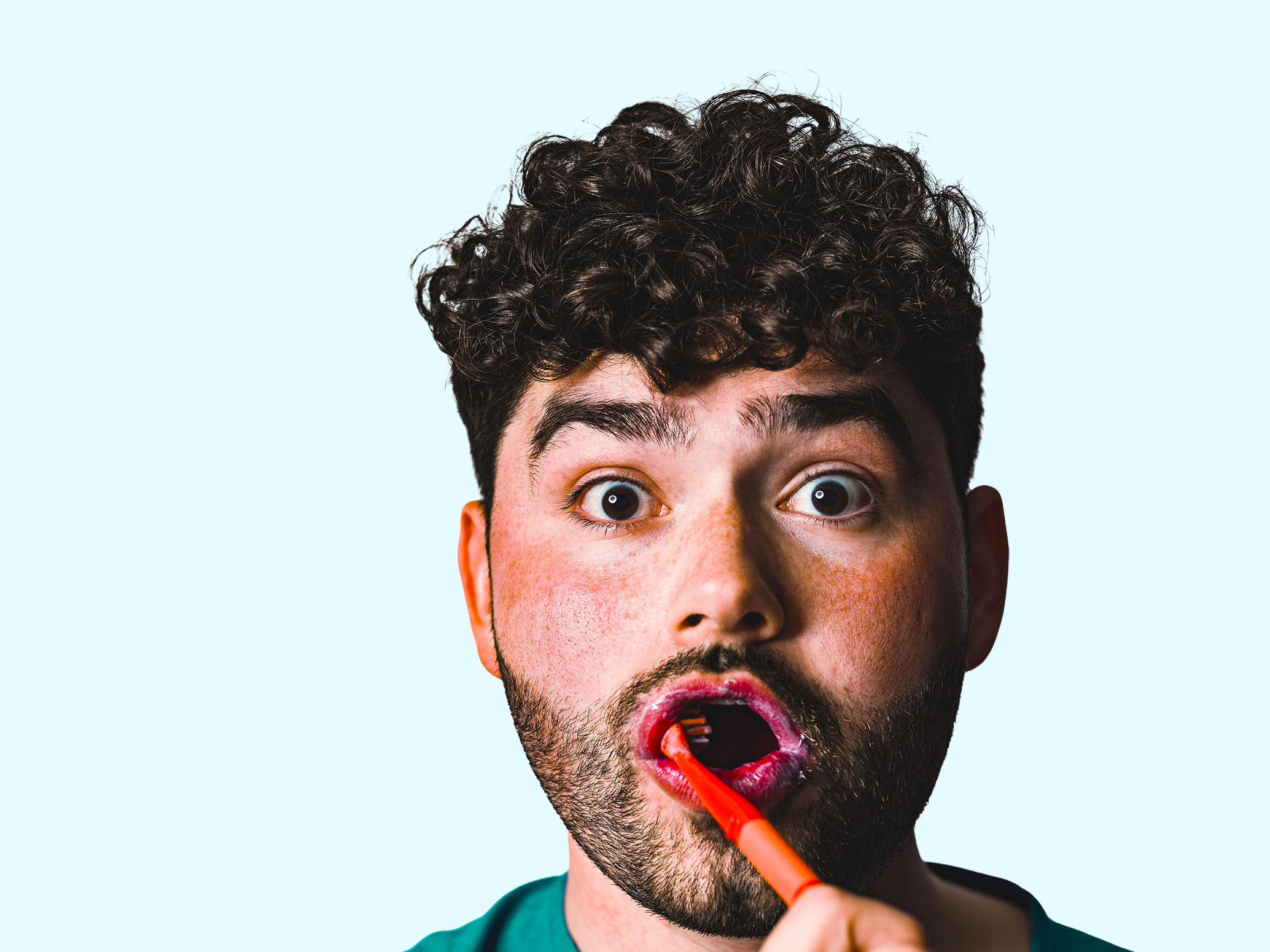 A man with curly hair and a beard is applying lipstick in front of a light blue background.