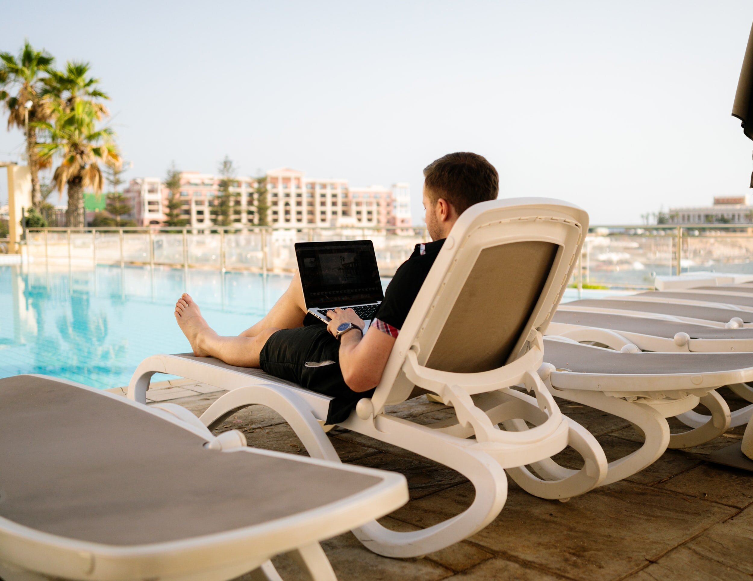 A picture of a financially independent man working from a laptop at the side of a pool.