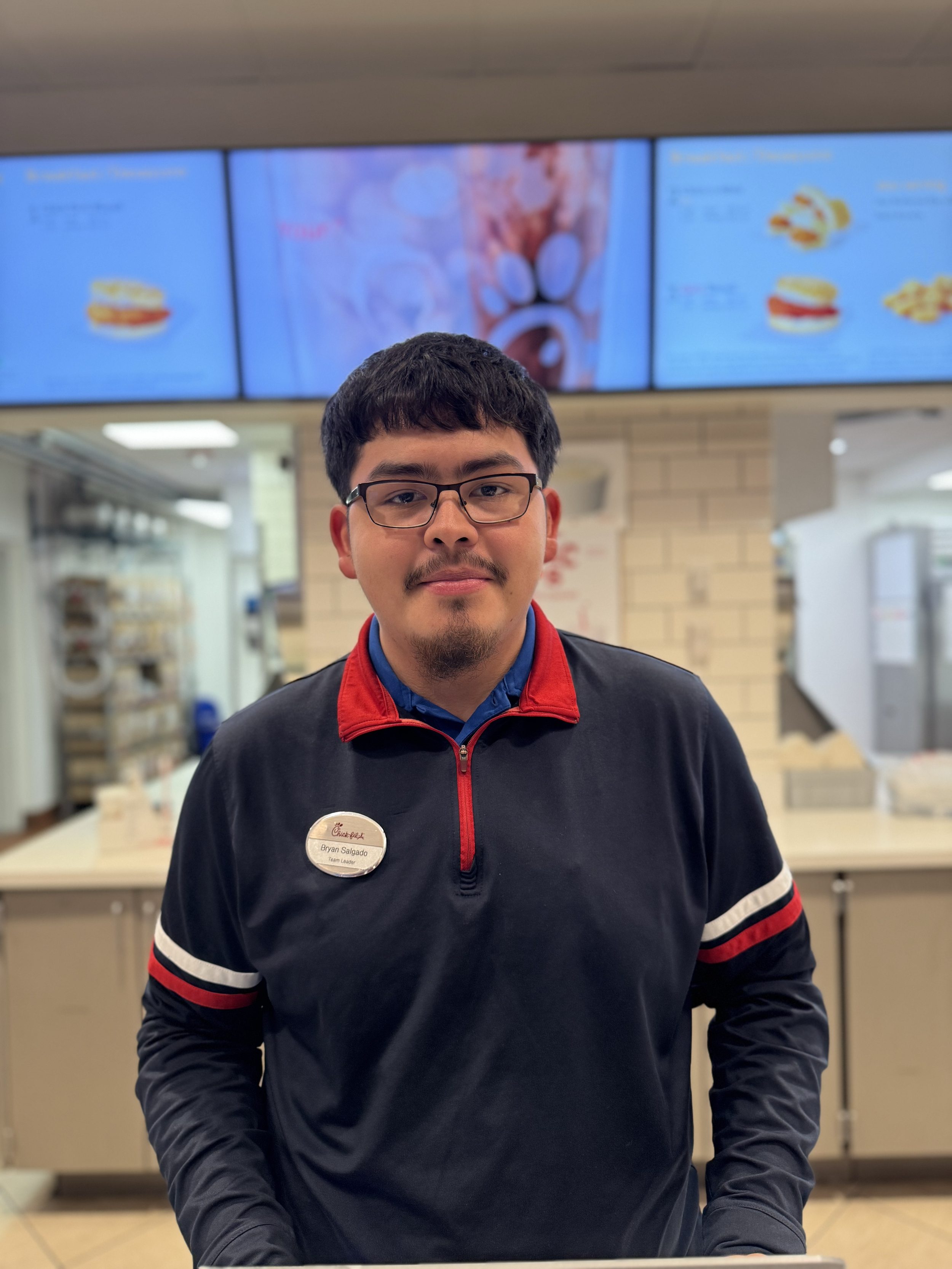 Male Chick-fil-A team member with glasses and a mustache, wearing a dark blue zip up and a name tag