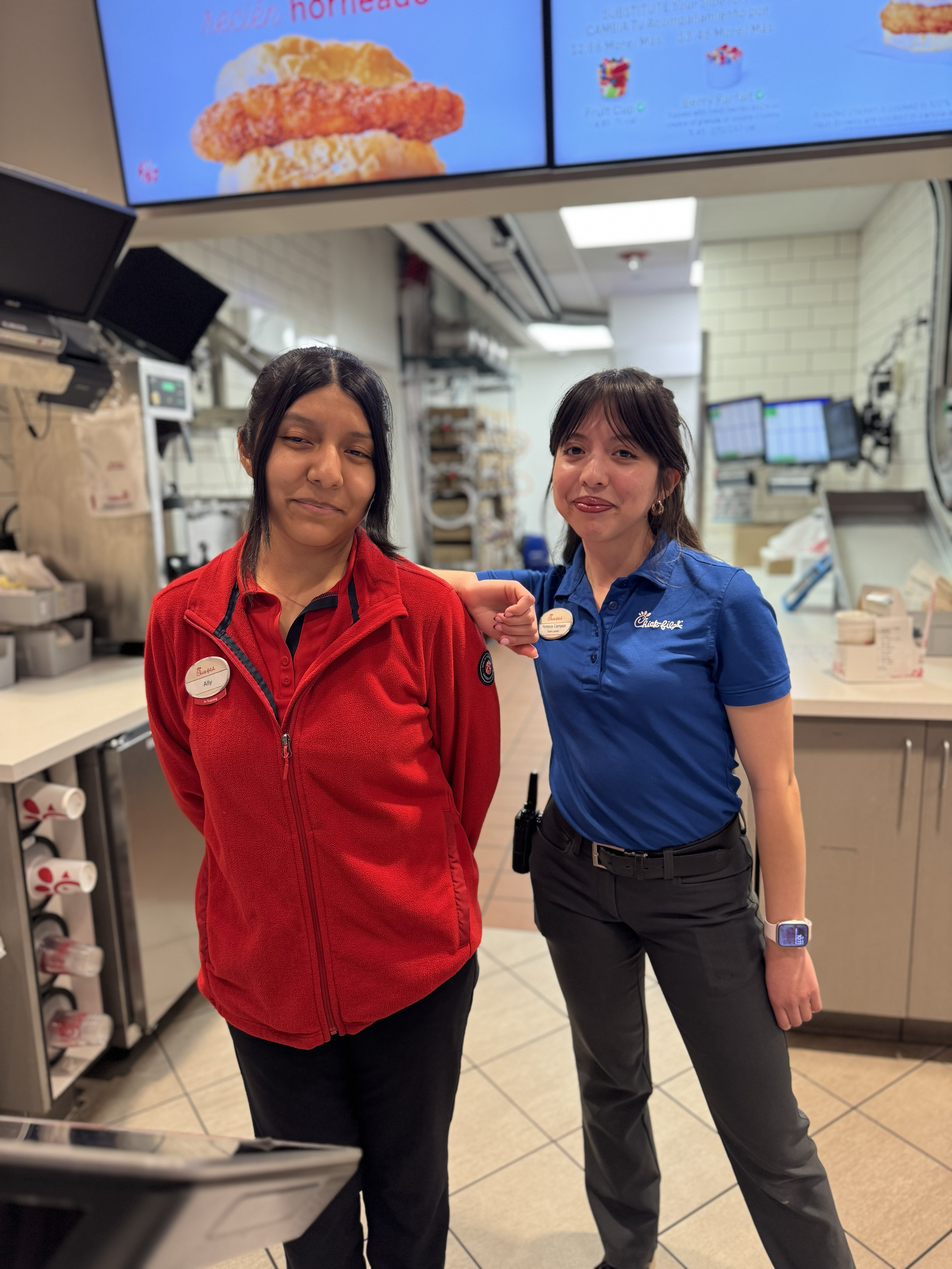 Two female Chick-fil-A team members, one in a red jacket and the other in a blue shirt. Both smiling at the camera.