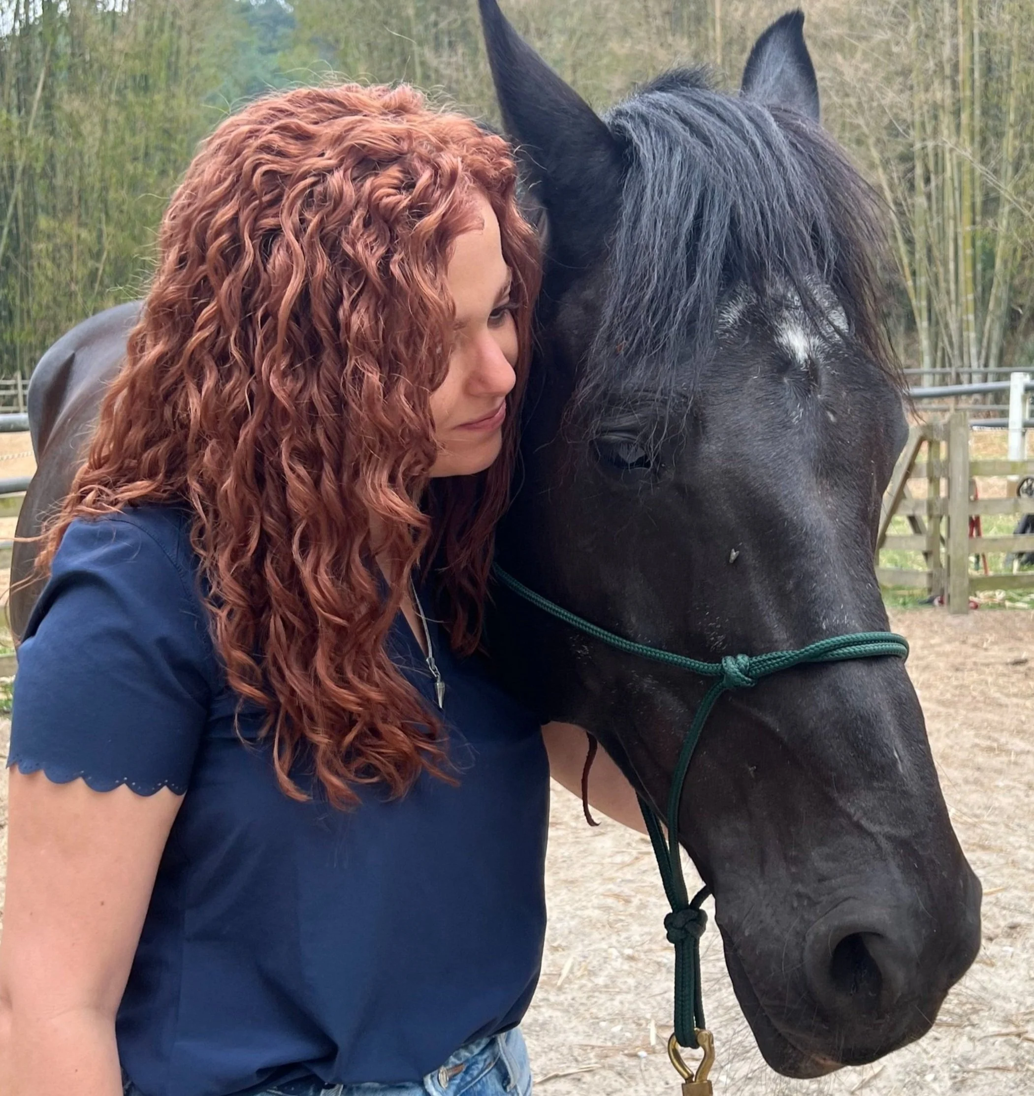 Una mujer con cabello rizado rojo oscuro apoyada en un caballo negro con melena larga, en un entorno de campo con cercas y árboles.