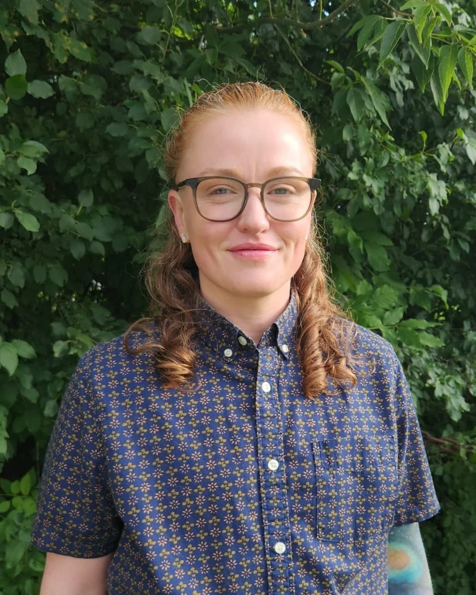 A person with pale skin and curly auburn hair, wearing glasses and a button-down shirt, standing in front of leaves.