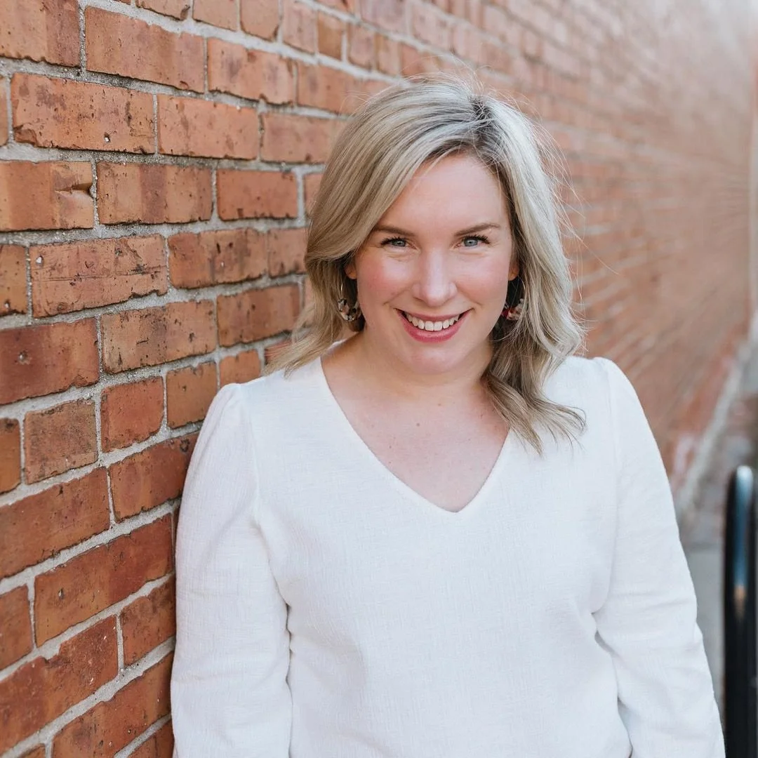 A woman with blonde hair, wearing a white top, smiling and leaning against a brick wall outdoors.