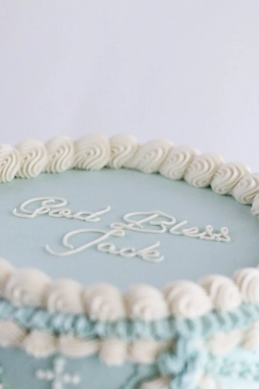 Close-up of the top of a light blue baptism cake with “God Bless Jack” piped in white script and surrounded by a white buttercream border.