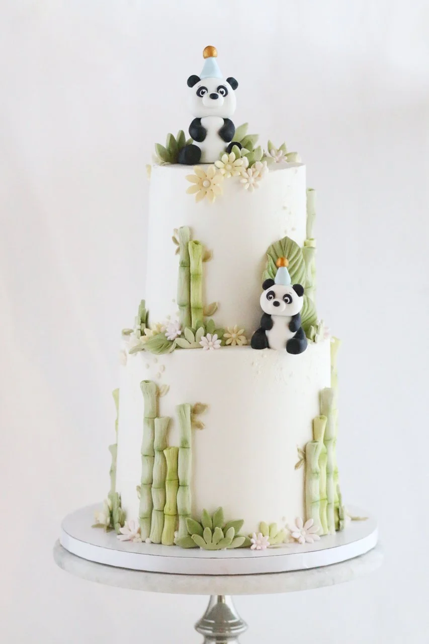 Two-tier white panda birthday cake decorated with two black-and-white pandas wearing blue birthday hats, green bamboo stalks, leaves, and small pastel flowers. Custom birthday cake by Rose Vanilla in Fairfax, Virginia.
