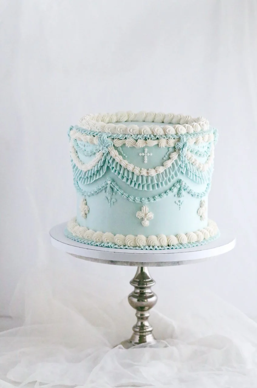 Light blue baptism cake with intricate white and blue buttercream piping, pearl cross details, and vintage-style swags, displayed on a silver cake stand.
