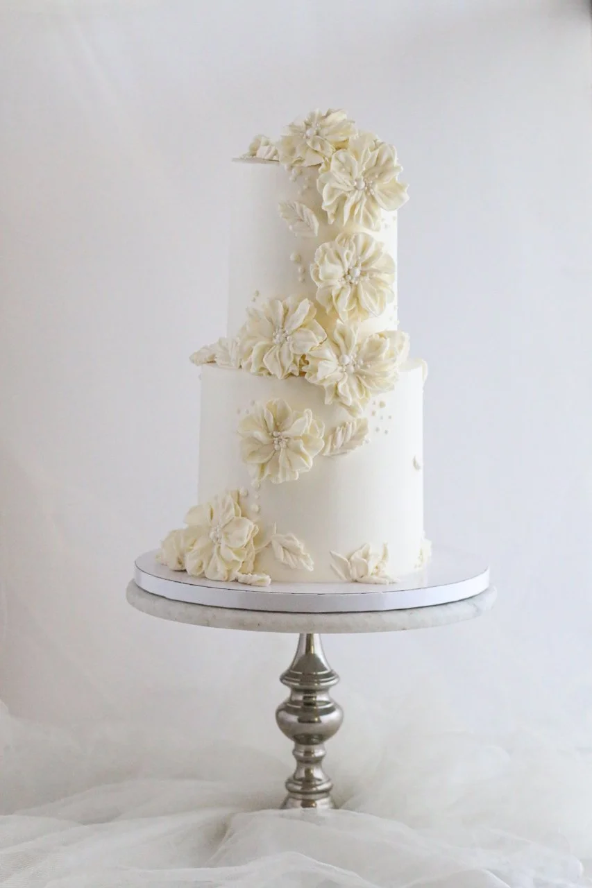 Two-tier ivory wedding cake with large hand-piped buttercream flowers, overlapping petals, delicate leaves, and small edible pearls on a silver cake stand.
