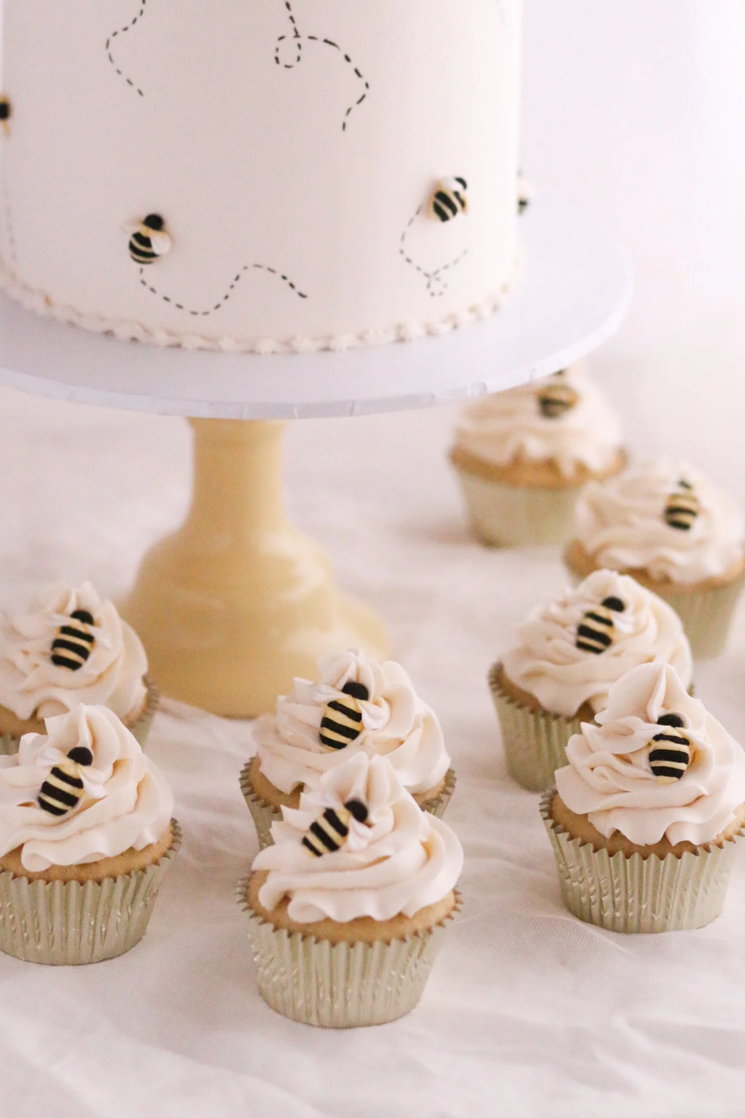 Minimalist white buttercream bee cake with dotted trails and matching swirl cupcakes topped with bee decorations
