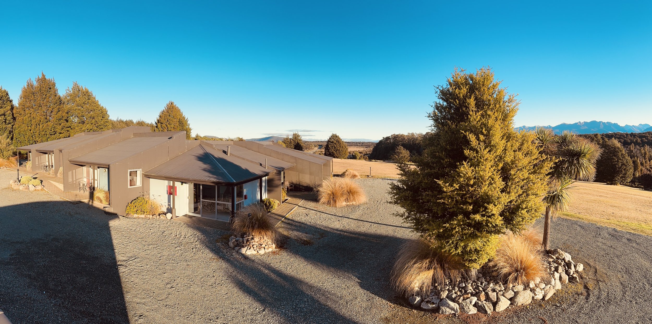 A modern building with a metal roof and glass doors, surrounded by desert landscaping, trees, and mountains in the distance under a clear blue sky.