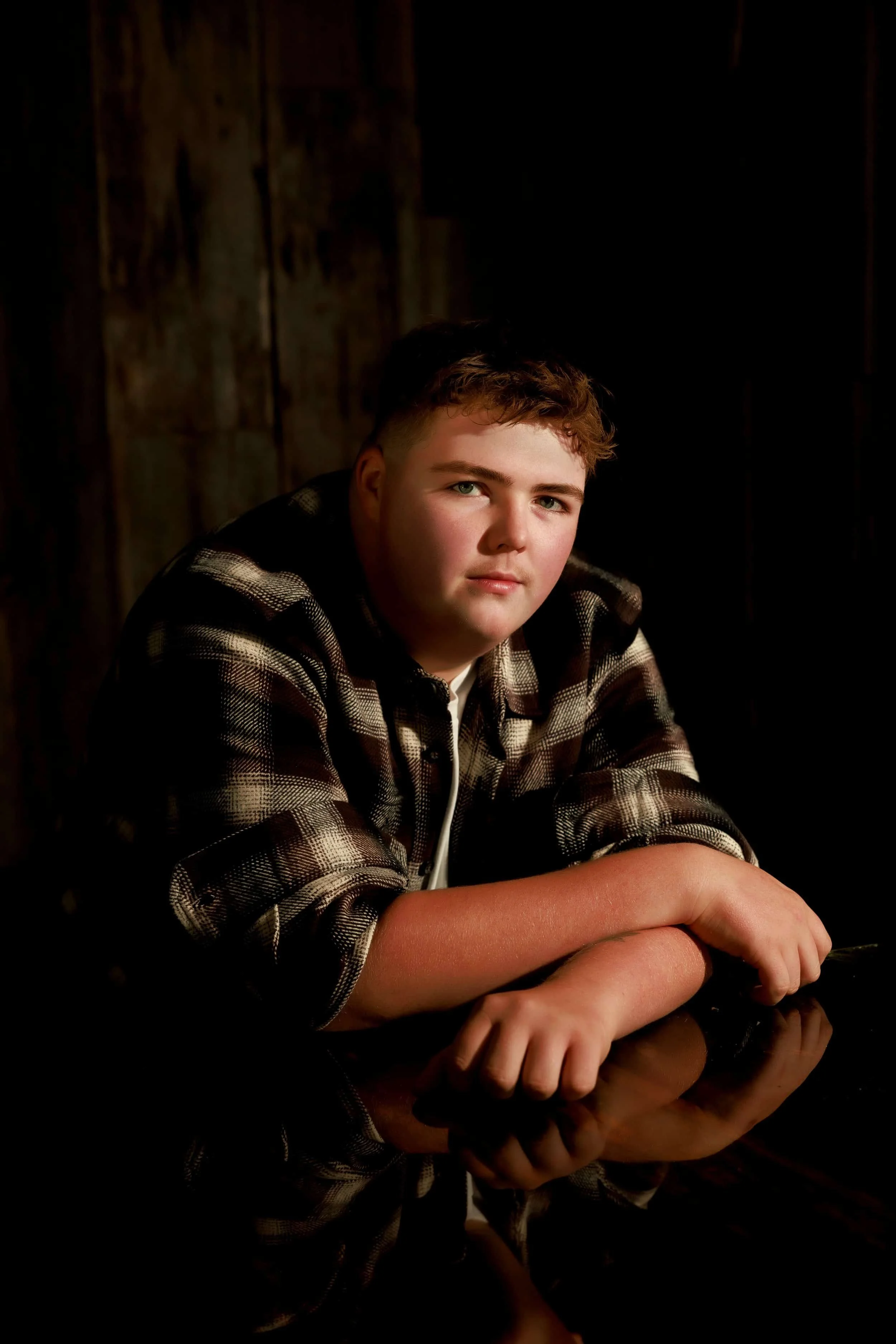 Senior photos near cameron mo, a boy wearing a plaid shirt, sitting at a reflective surface with a dark background.
