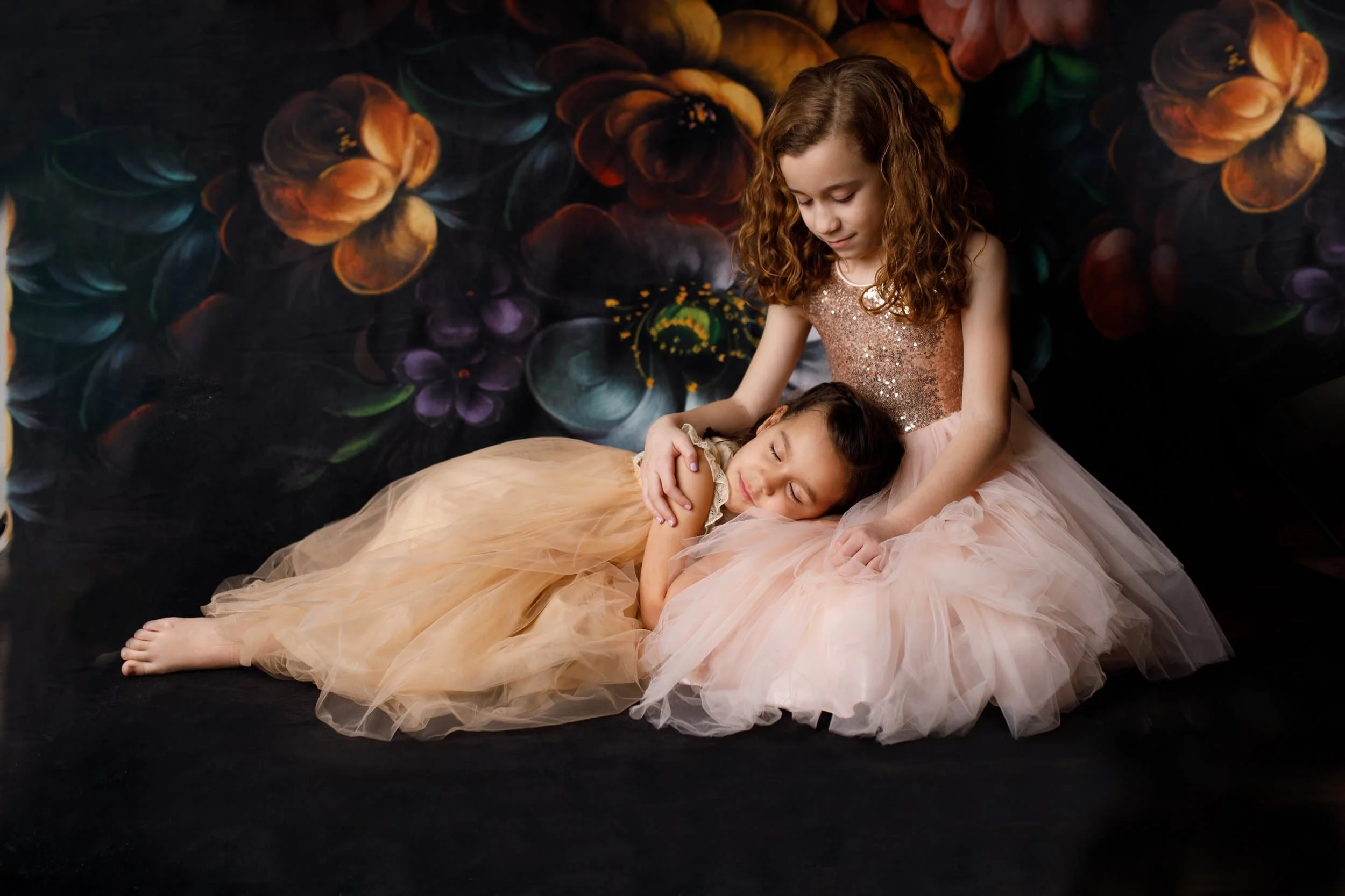 Two young girls in fancy dresses, one lying down with eyes closed and the other sitting beside her, comforting her in front of a dark floral backdrop. In Cameron MO