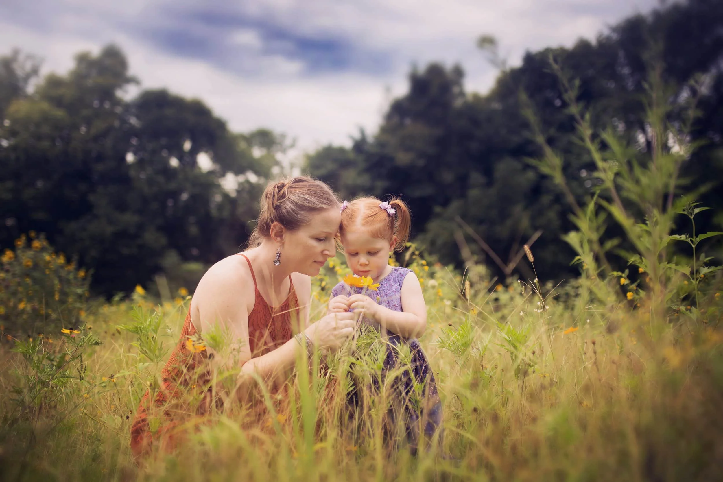 A woman and a young girl in a grassy field holding a yellow flower together, with trees and a cloudy sky in the background. Near Cameron MO