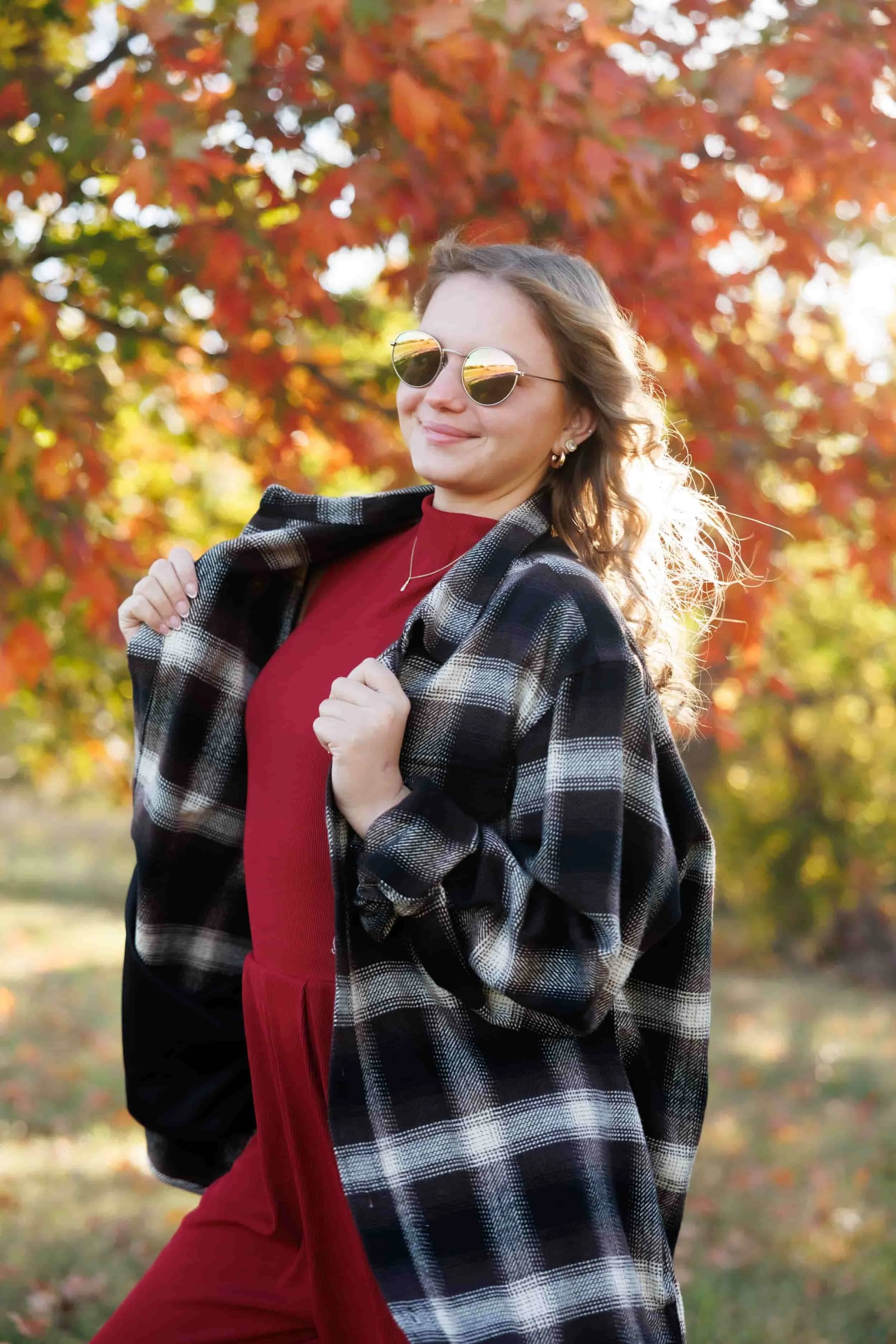 A woman with wavy hair wearing sunglasses, a red dress, and a black and white checkered jacket, standing outdoors with fall-colored trees in the background.