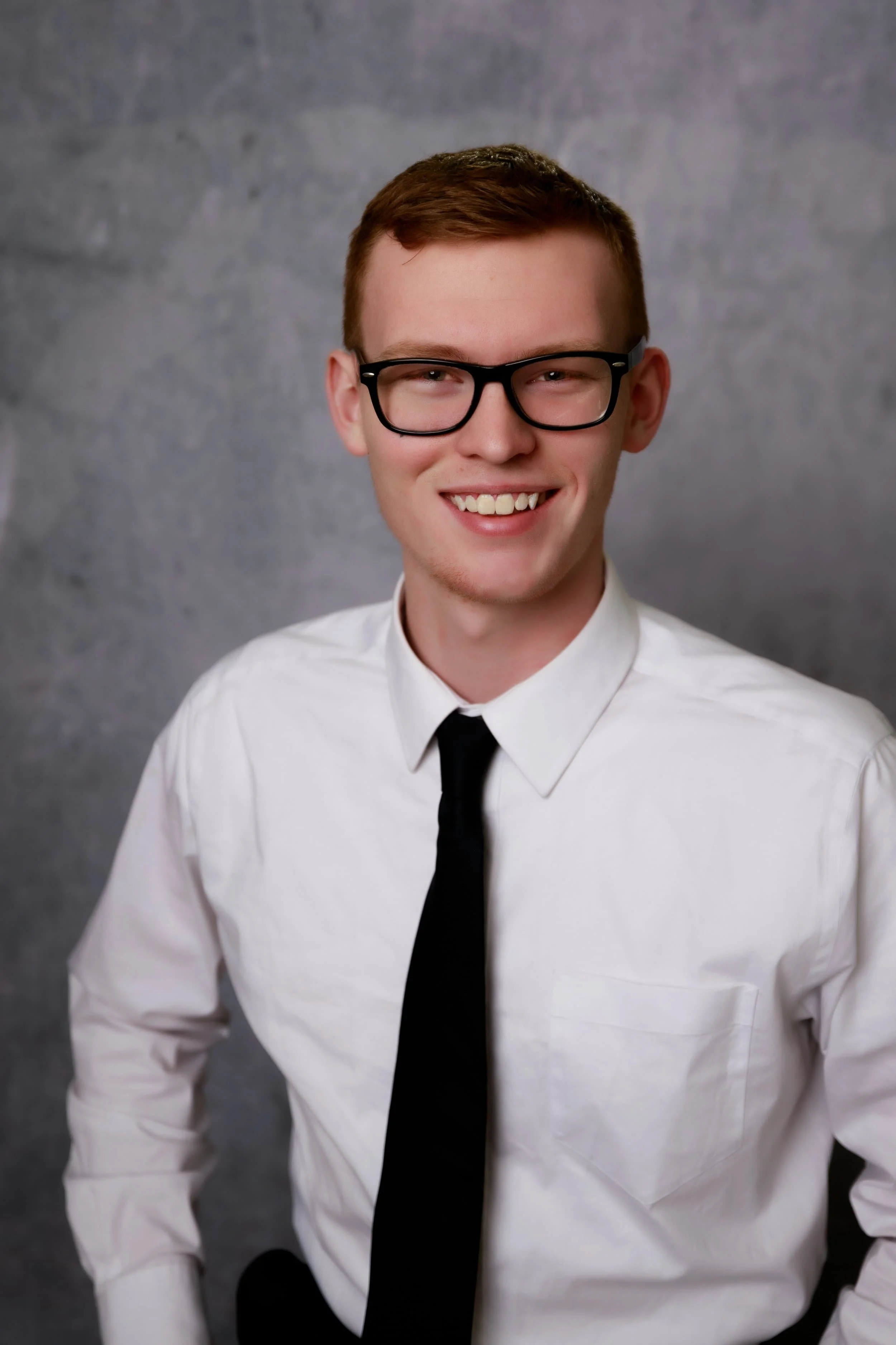 Young man with glasses, wearing a white shirt and black tie, smiling, posing against a gray background for senior photos in cameron mo