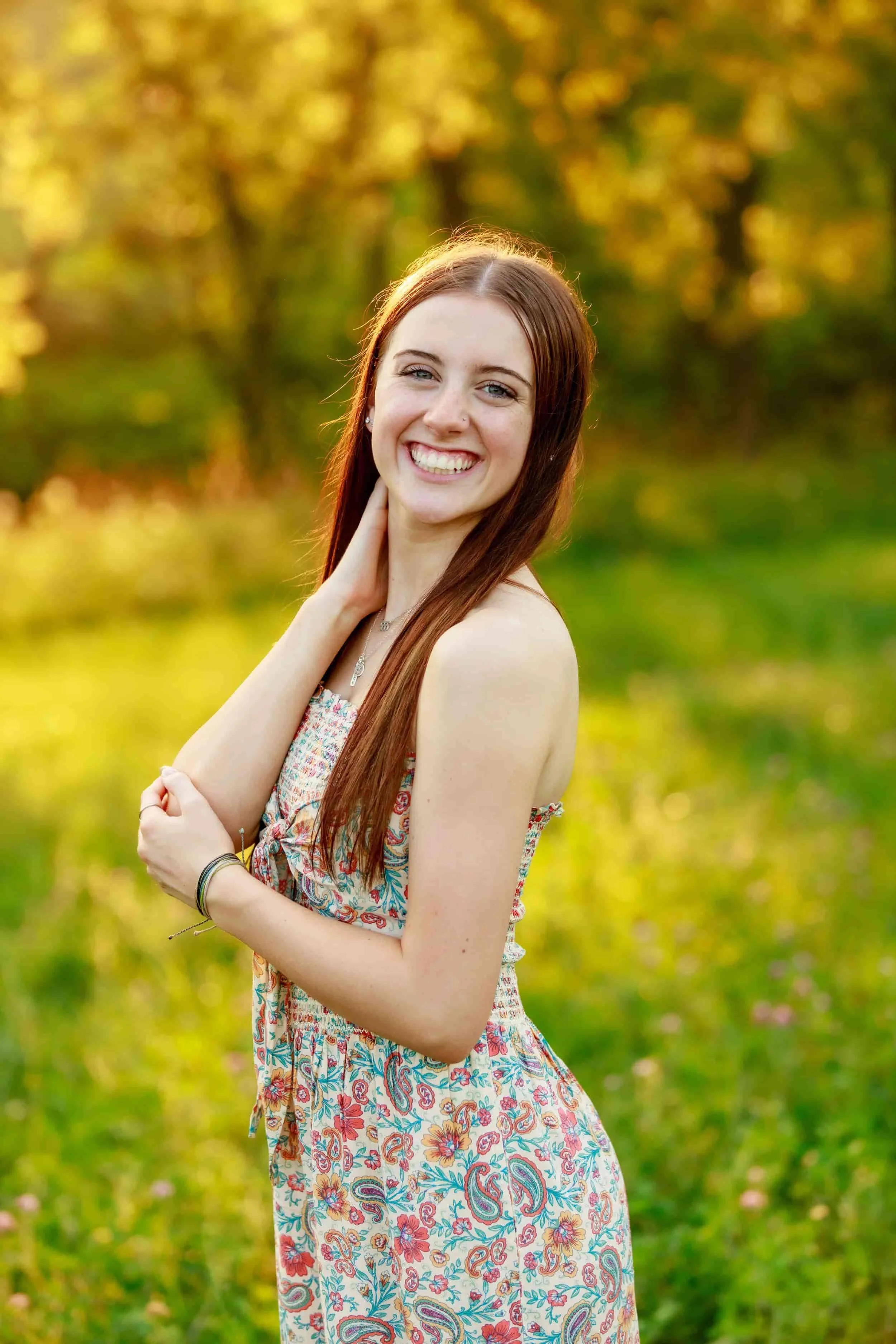 A young woman with long red hair, smiling with teeth showing, standing outdoors in a park during golden hour. She is wearing a strapless, floral dress and has her right hand gently touching her neck, with greenery and trees in the blurred background.