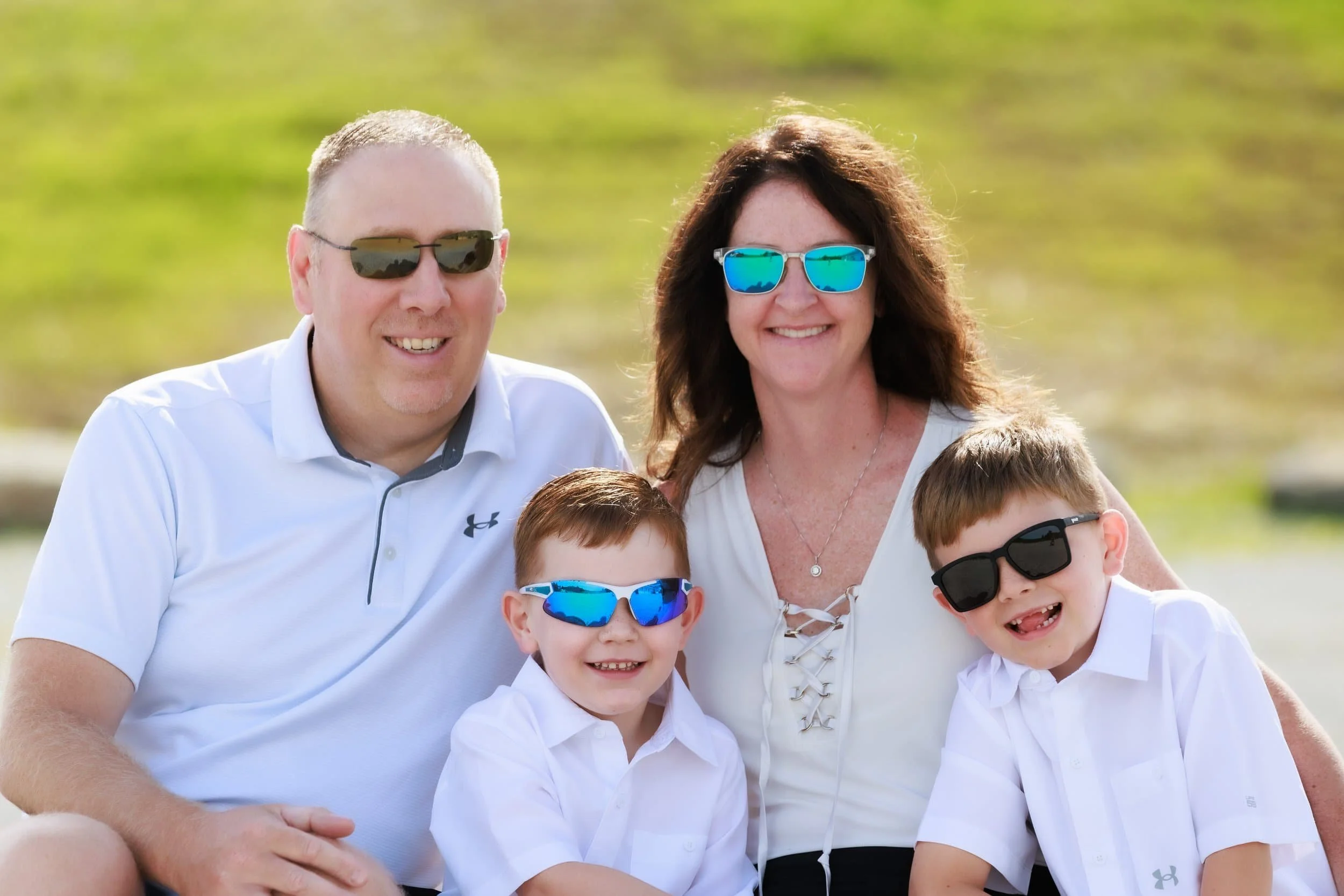 Family of four Near Cameron MO, two adults and two children, smiling and wearing sunglasses outdoors on a sunny day. The adults are wearing white tops, and the children are in white shirts.