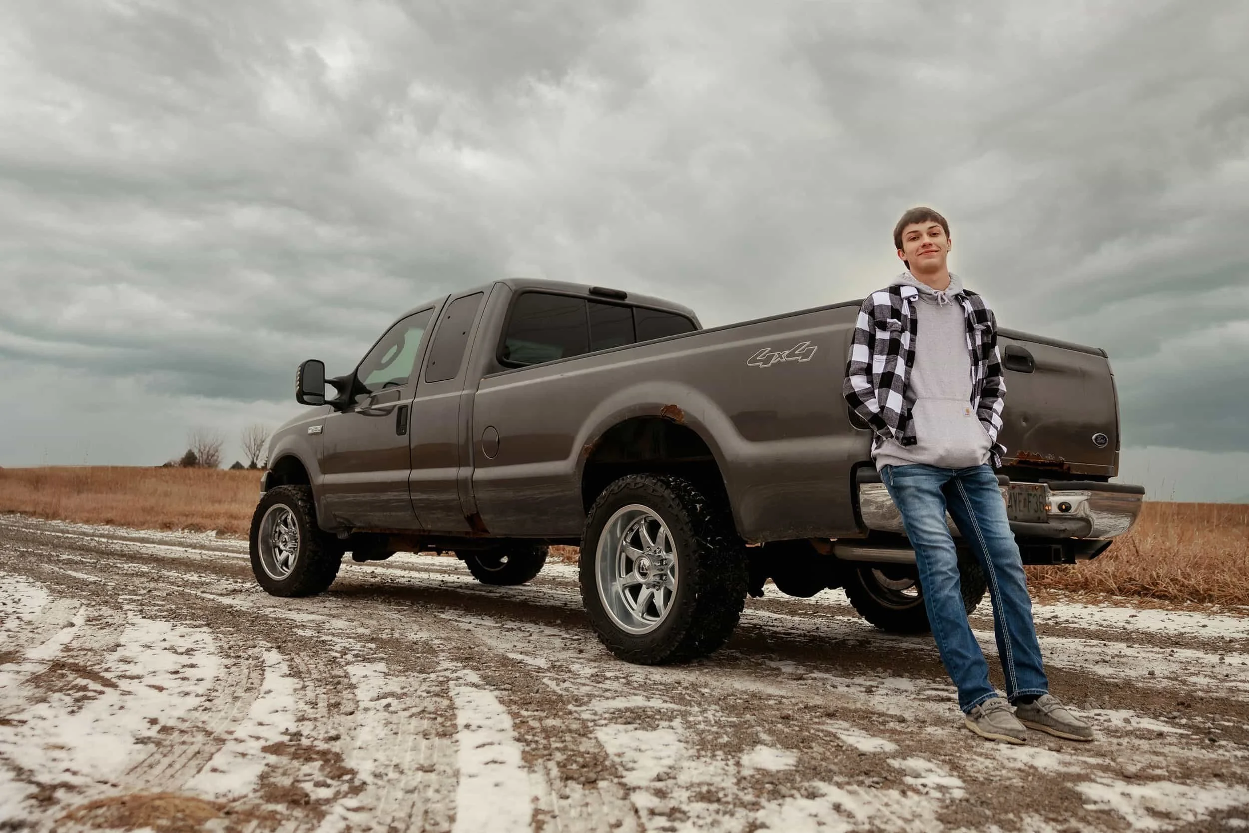 Young man standing outside on a dirt road near saint joseph mo for senior photos with patches of snow, leaning against a dark gray Ford pickup truck with a rusty rear corner, under a cloudy sky.