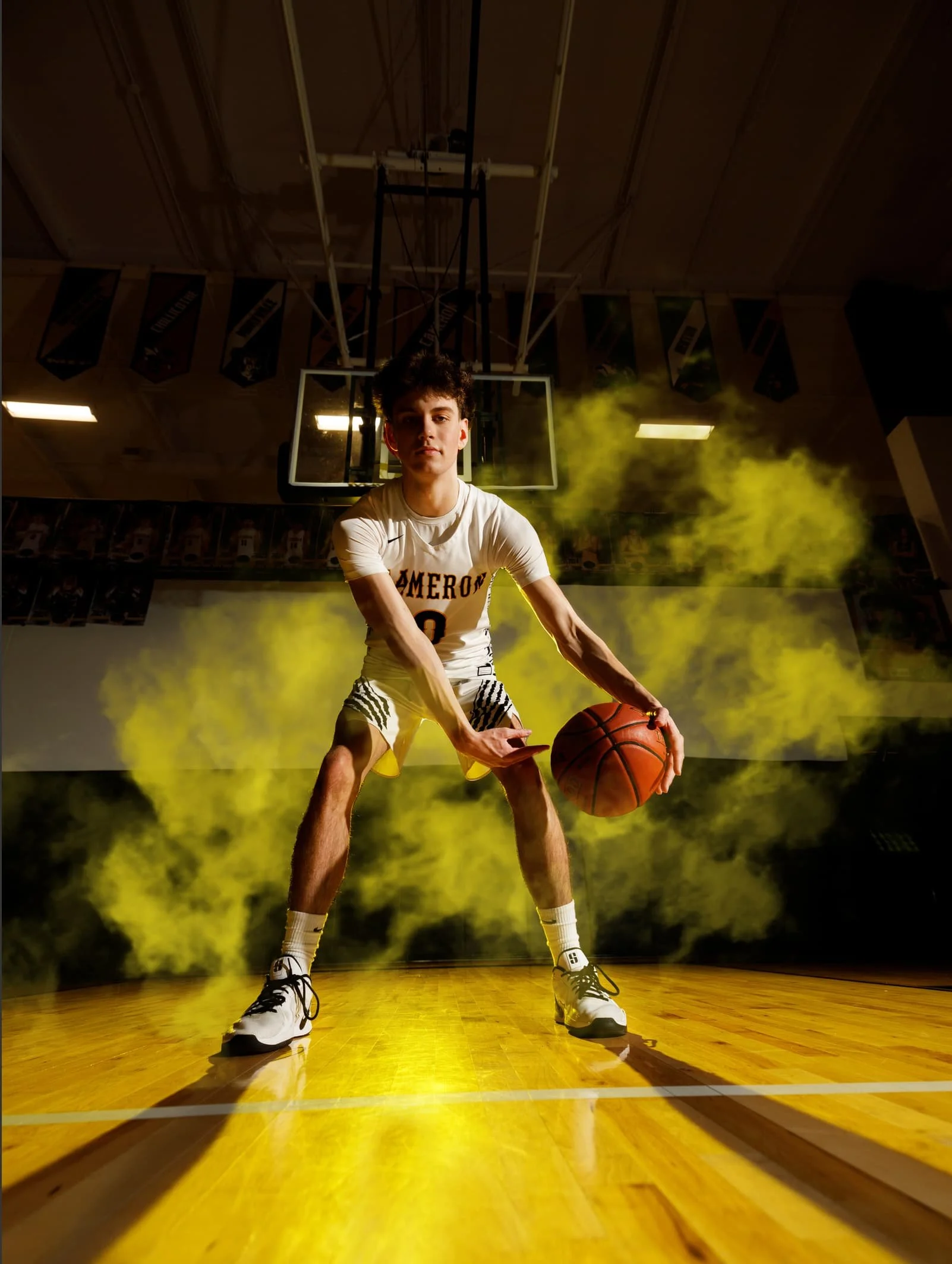 A young male basketball player in a white uniform with black and yellow accents, holding a basketball in a gymnasium with a basketball hoop behind him. Yellow smoke surrounds him, with dramatic lighting on a polished wooden floor.
