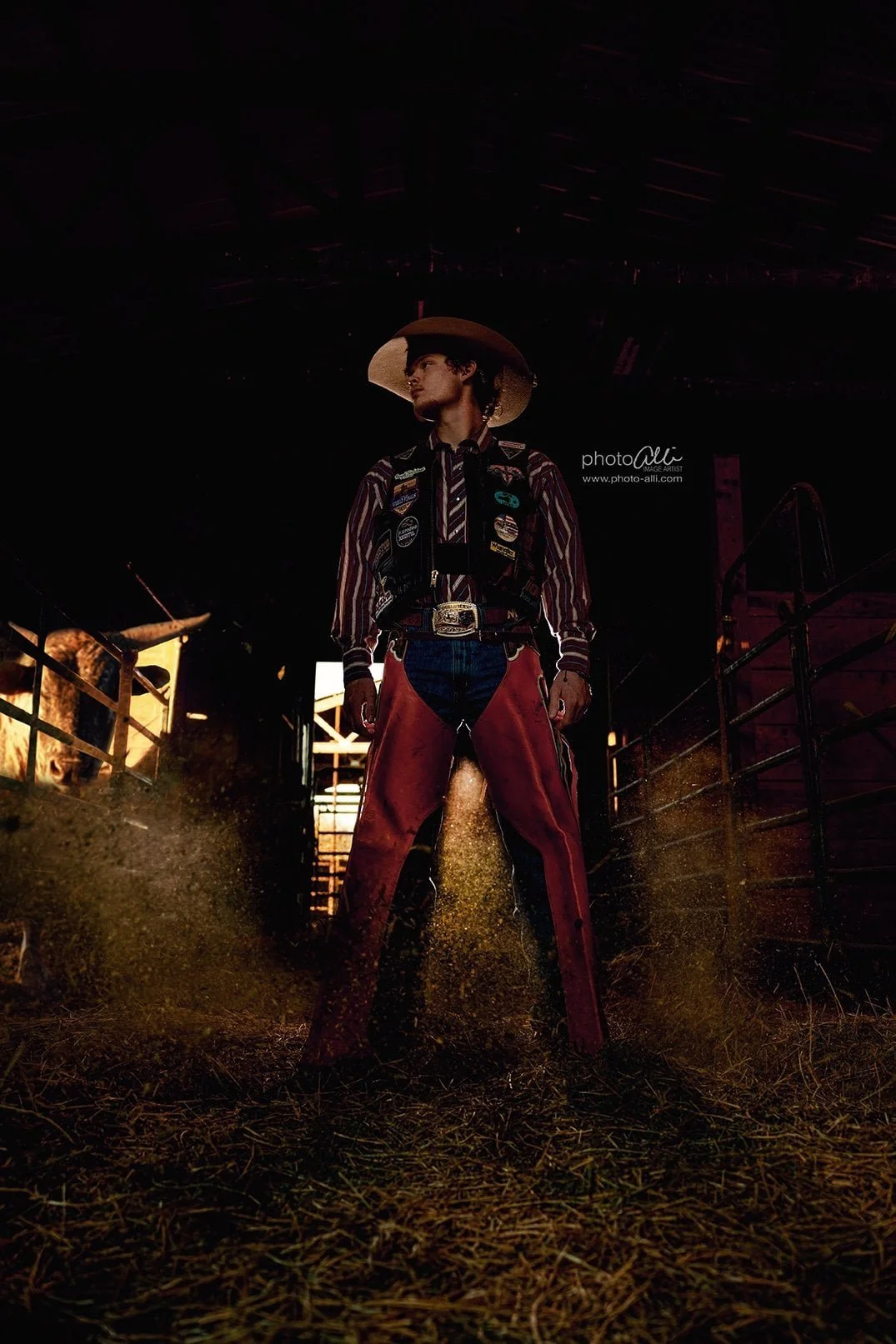 in cameron mo a Person dressed as a cowboy standing in a barn, with a cow visible on the left side, wearing a vest with patches, a striped shirt, and red chaps, with sunlight coming through the barn.