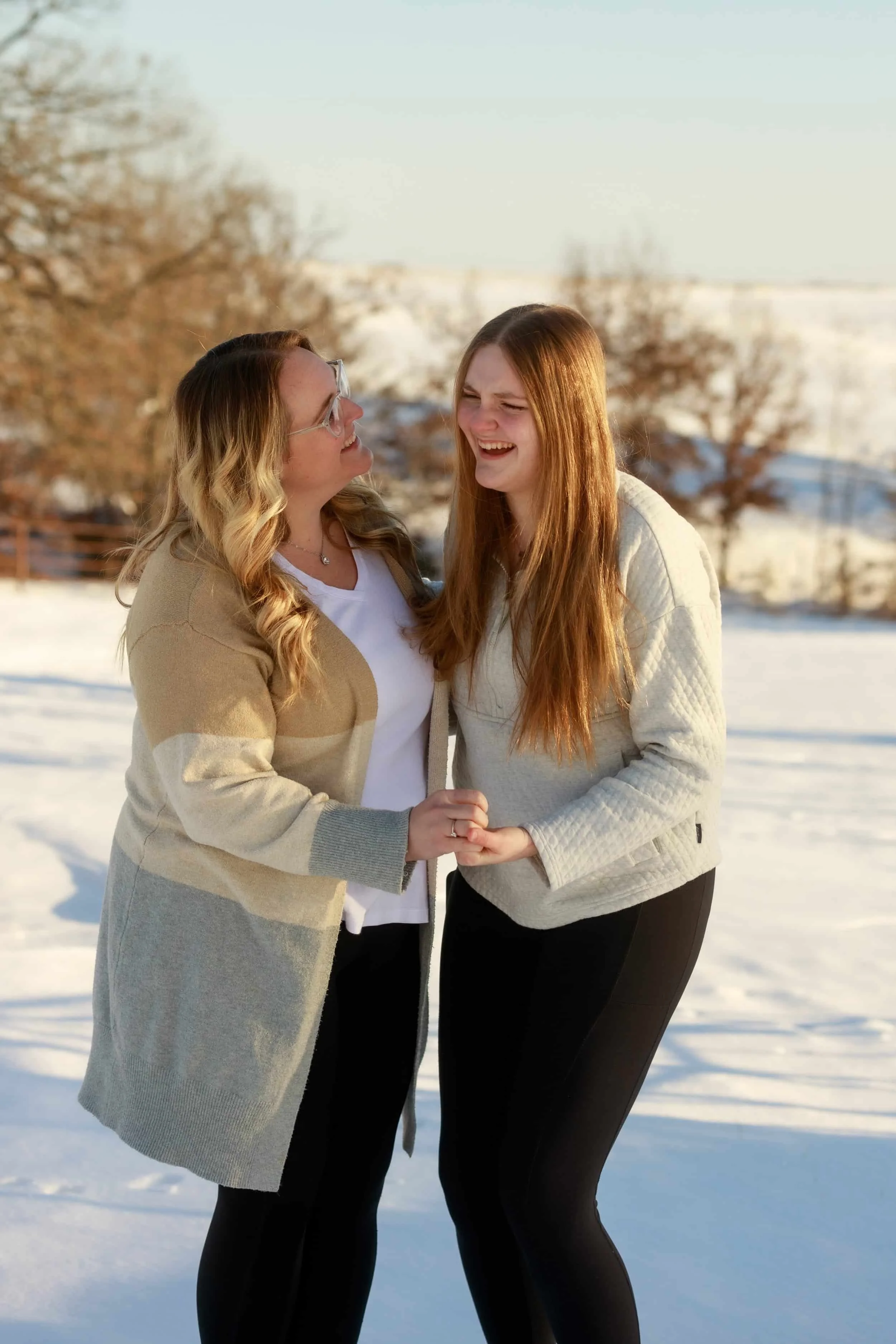 Two women Near Cameron MO laughing and holding hands in a snowy outdoor setting with snow-covered trees and a wooden fence in the background.