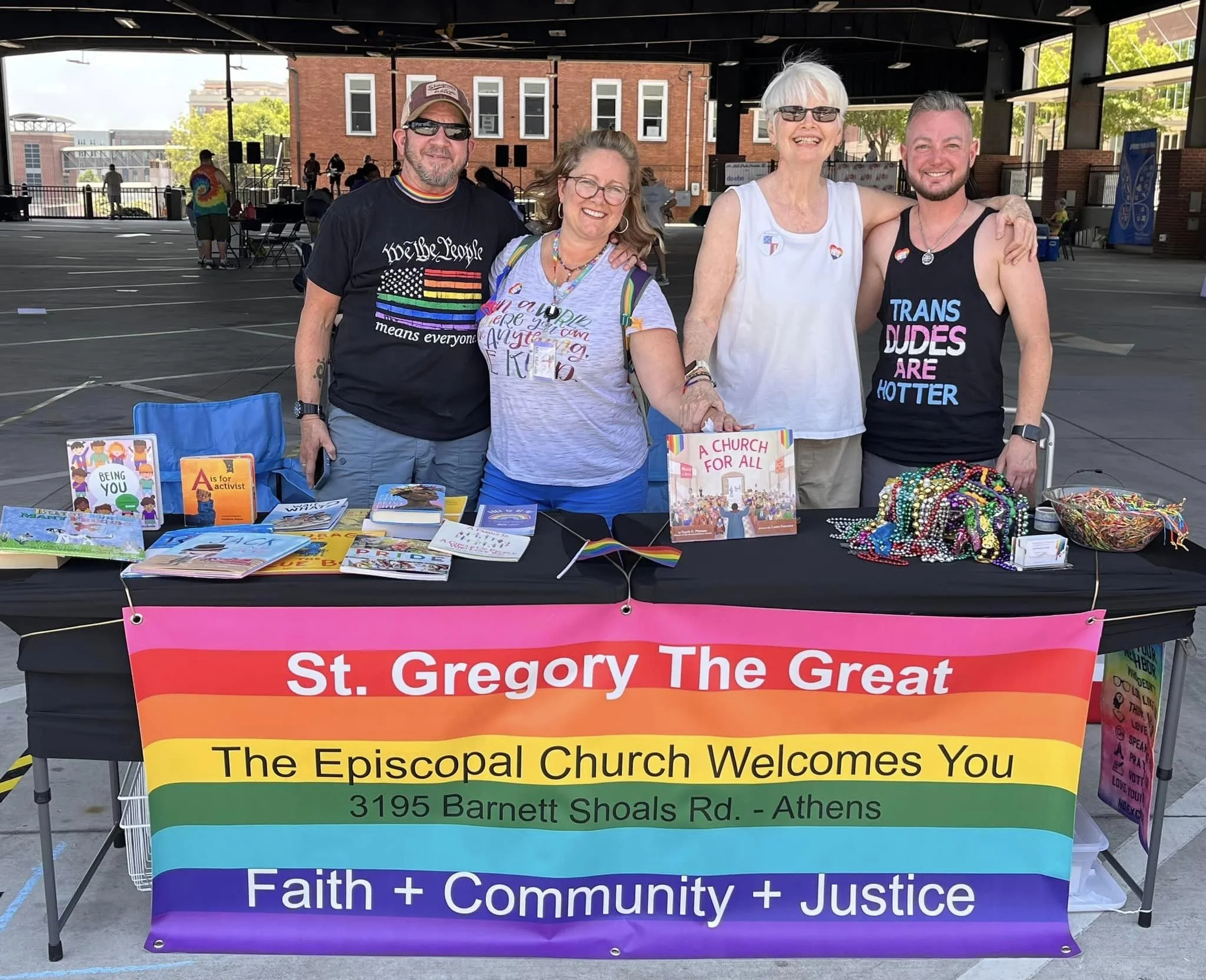 Two men and two women wearing LGBTQA+ affirming t-shirts, in a booth displaying beads, literature, Pride flags, and other giveaway items
