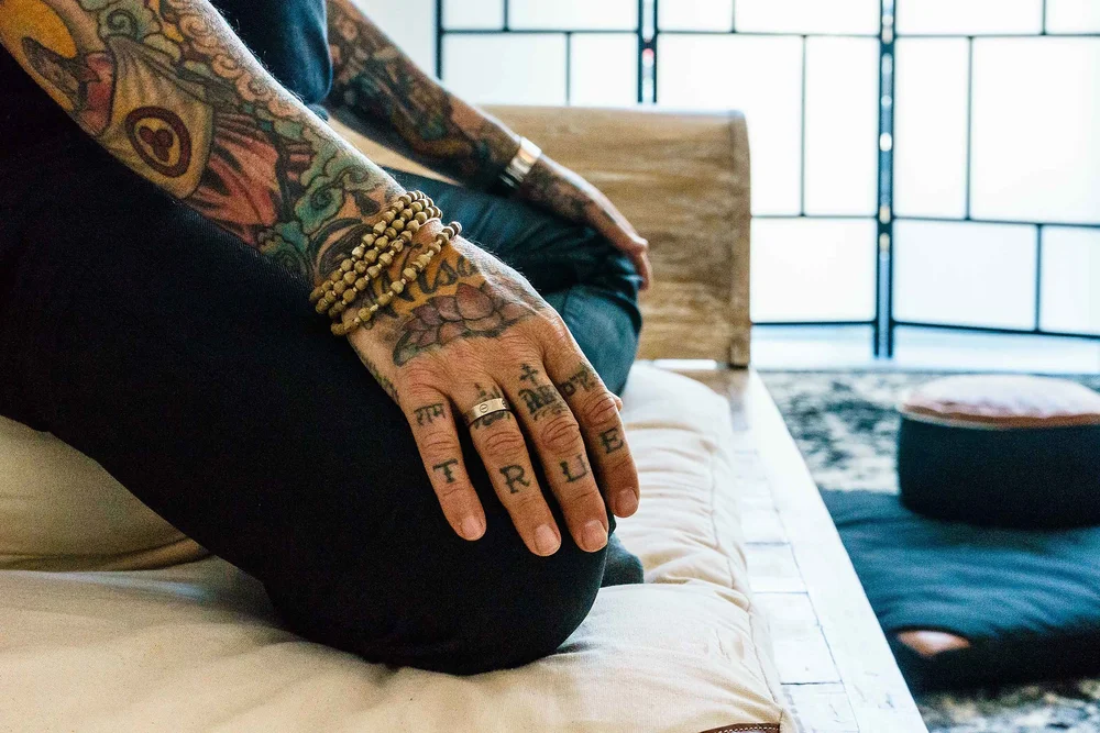 Close-up of a sitter's tattooed hands resting on the knees, cross-legged on a cushion in the hall