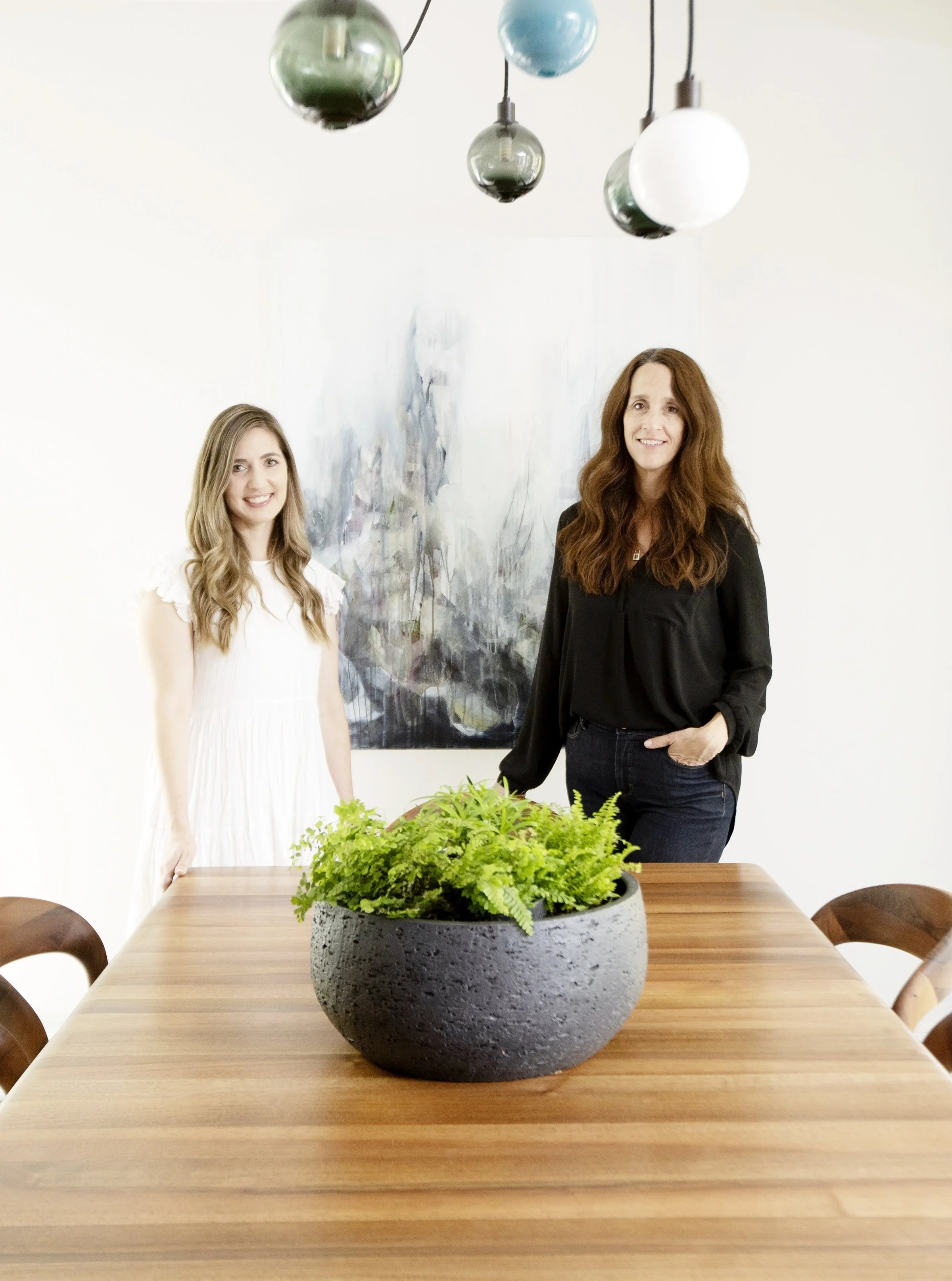 Trish Knight and Nicole Varga stand behind a dining table with a large potted green plant, in a room with modern lighting and abstract artwork on the wall.