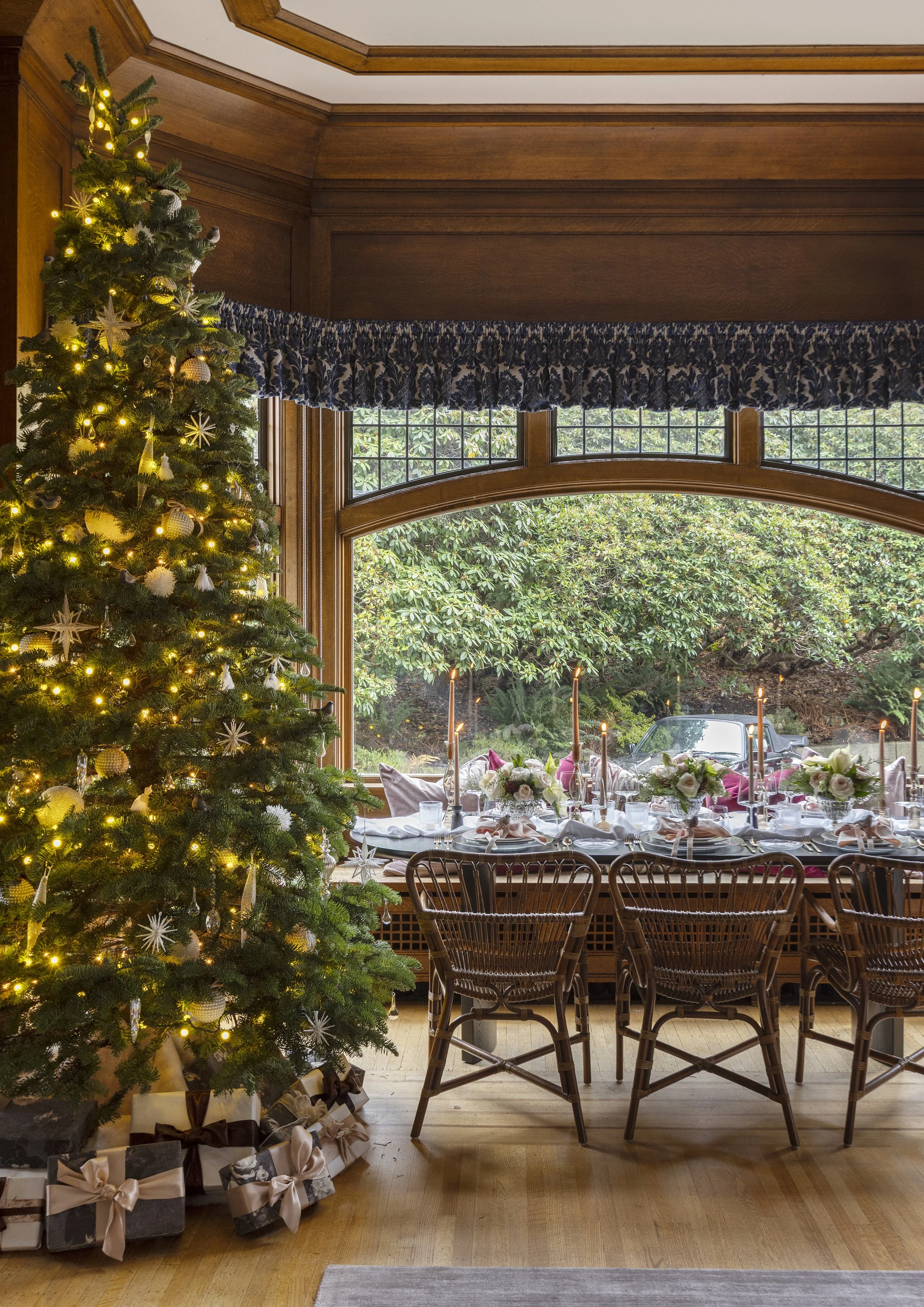 A decorated Christmas tree with lights and presents underneath in a dining room with a large window and a holiday table setting.