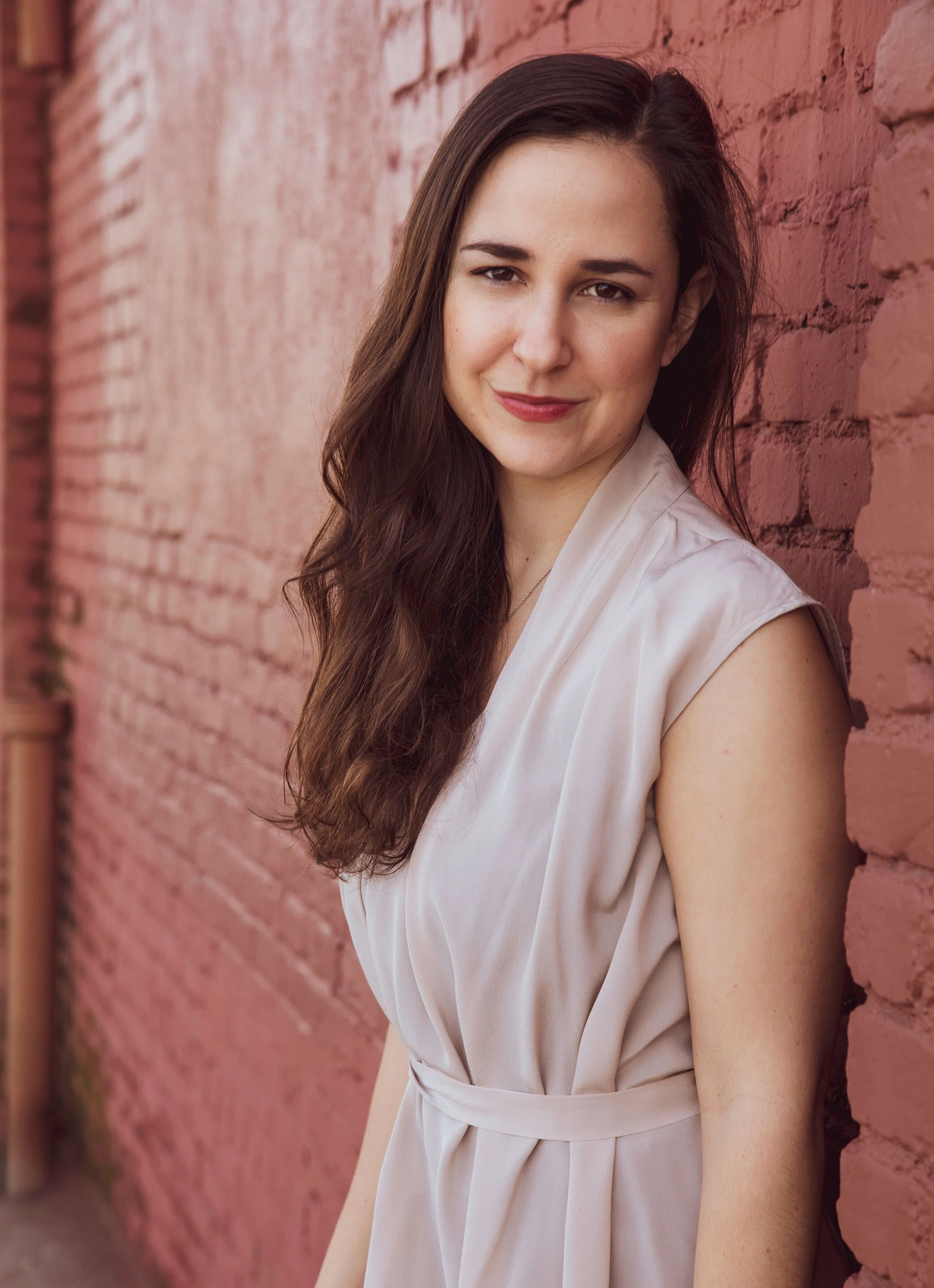 The torso and face of a person with long dark hair, wearing a light-colored dress, standing against a pink brick wall, looking at the camera with a slight smile.