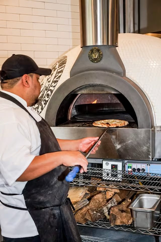 Chef placing a pizza into a wood-fired oven in a restaurant kitchen.