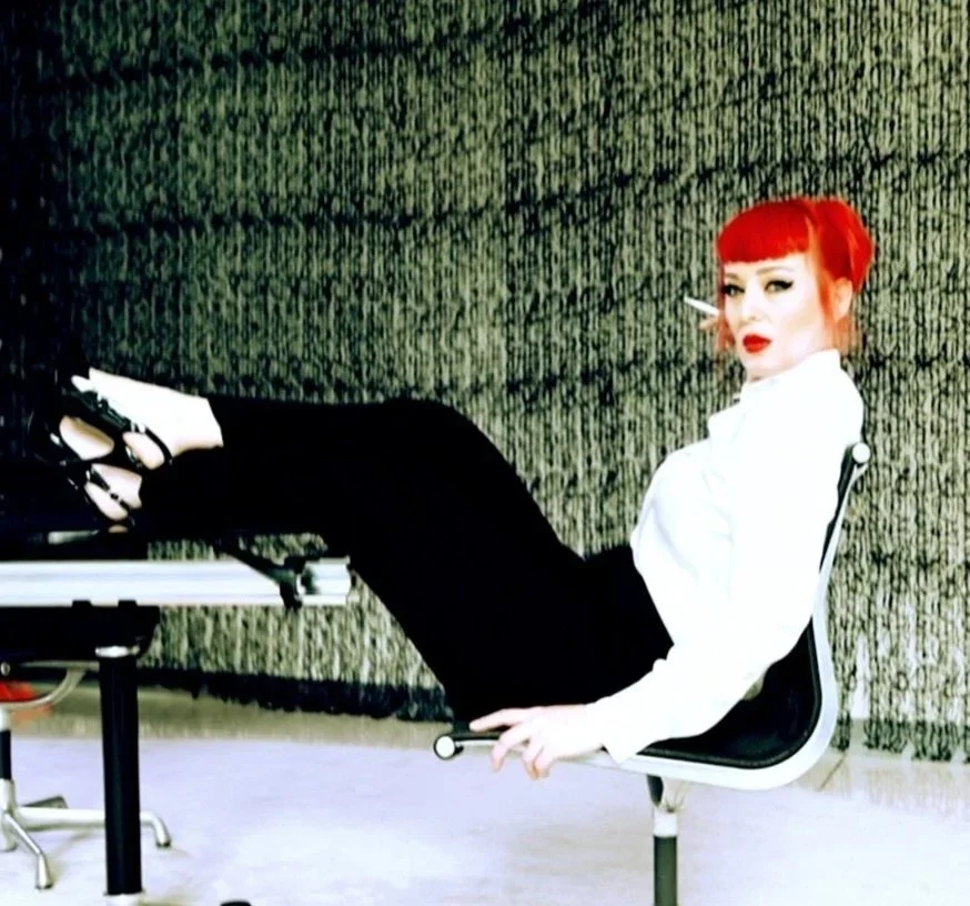 Mistress Zoe Demure, ready to teach a BDSM Workshop with feet up on her desk looking Dominant