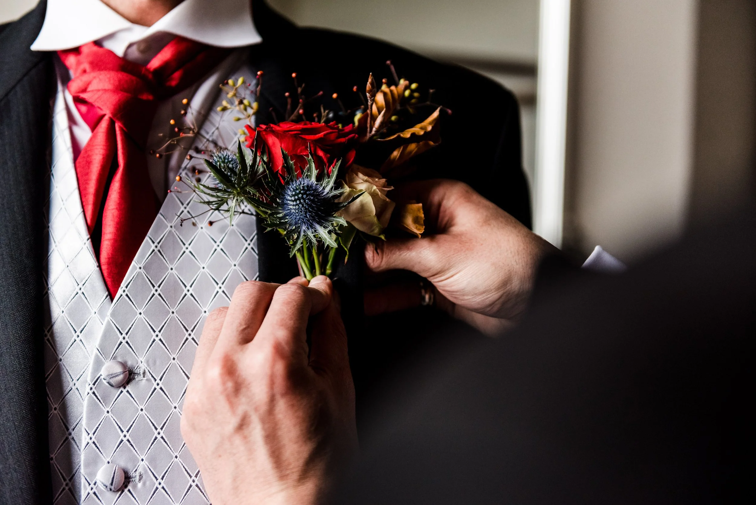 A grooms button hole containing a red rose, pink spray rose, eryngium, copper beech.