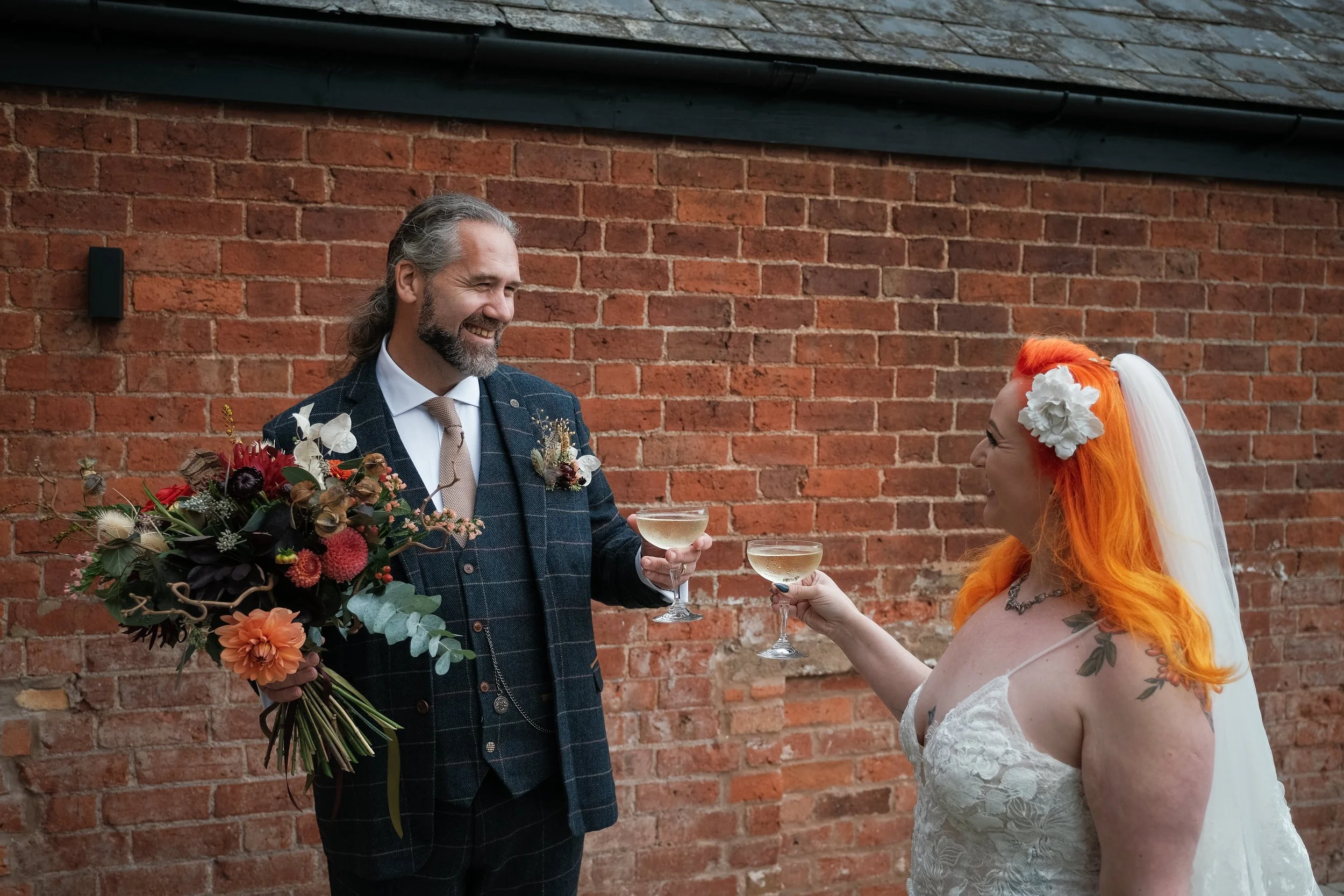 A bride and groom drinking champagne, the groom is wearing a pocket meadow and is holding the bouquet which includes british dahlias.