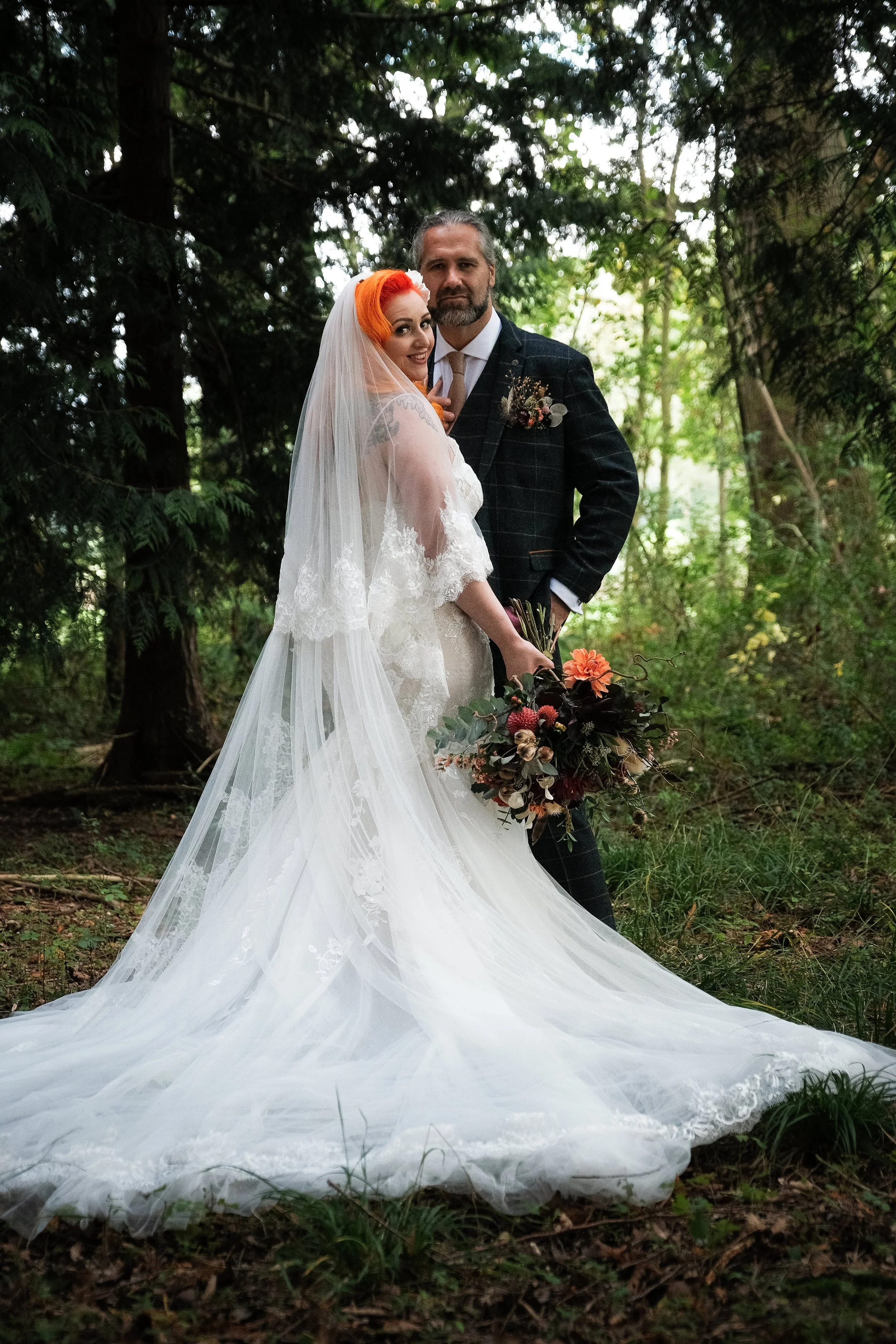 A bride and groom in the woodland setting of their wedding. The bride is holding a wild boho style bouquet in autumnal colours, including Dahlias, the groom is wearing a pocket meadow instead of a pocket square.