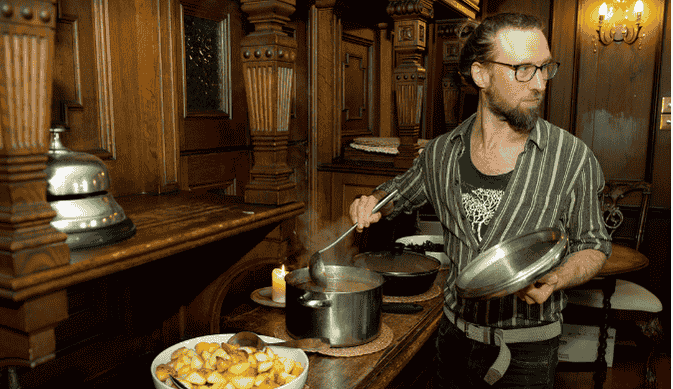 A man with glasses, a striped shirt, and a beard cooking in a wooden-paneled kitchen, holding a lid in his left hand and stirring a pot on the stove with a spatula in his right hand, with a bowl of cooked potatoes on the counter.
