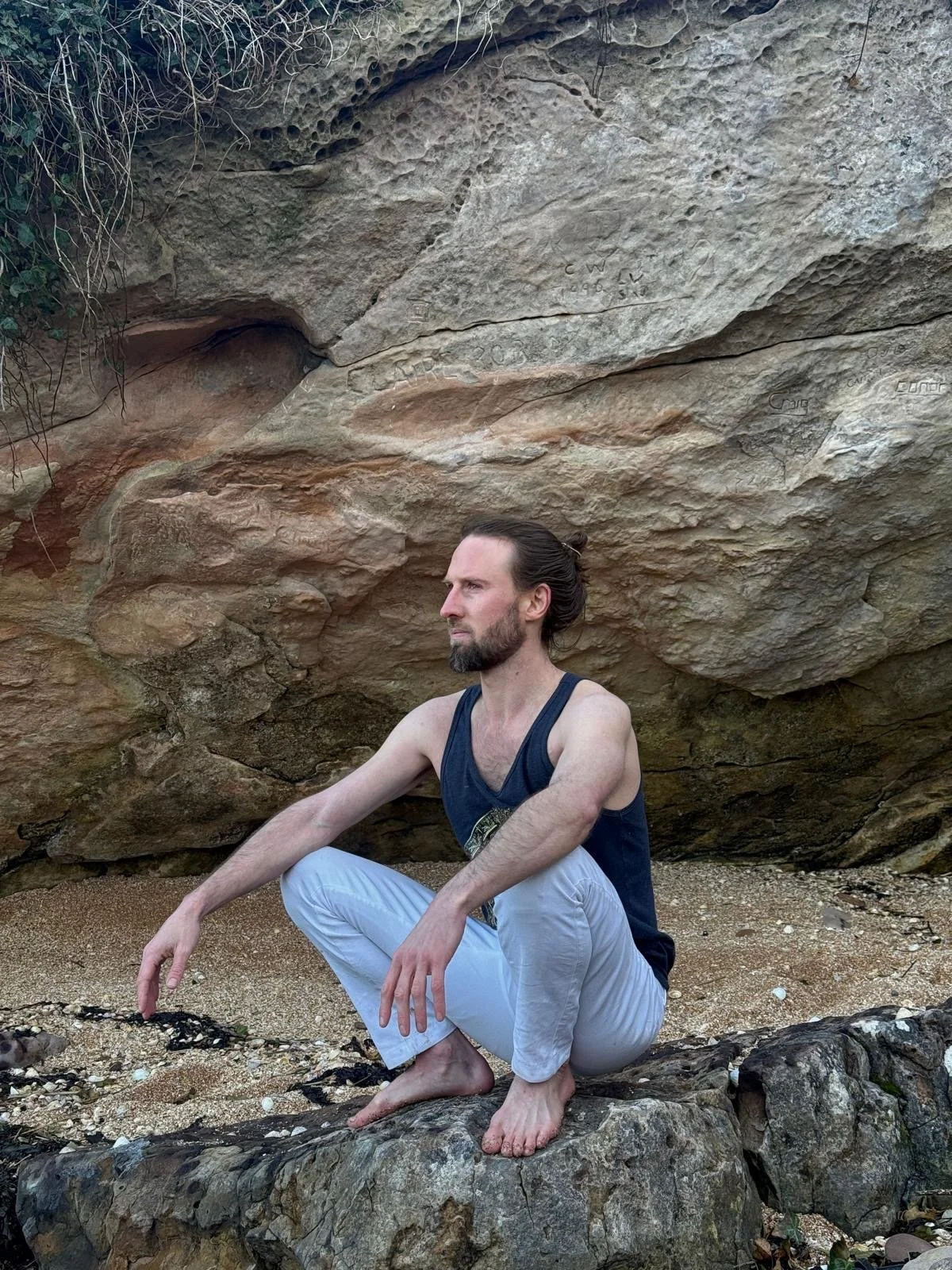 A man with long hair tied back, with a beard, dressed in a black sleeveless shirt and white pants, is crouching barefoot on a rock in front of a large cave-like rock formation. He appears to be meditating or in a contemplative pose near a sandy area with some scattered black sticks or branches.