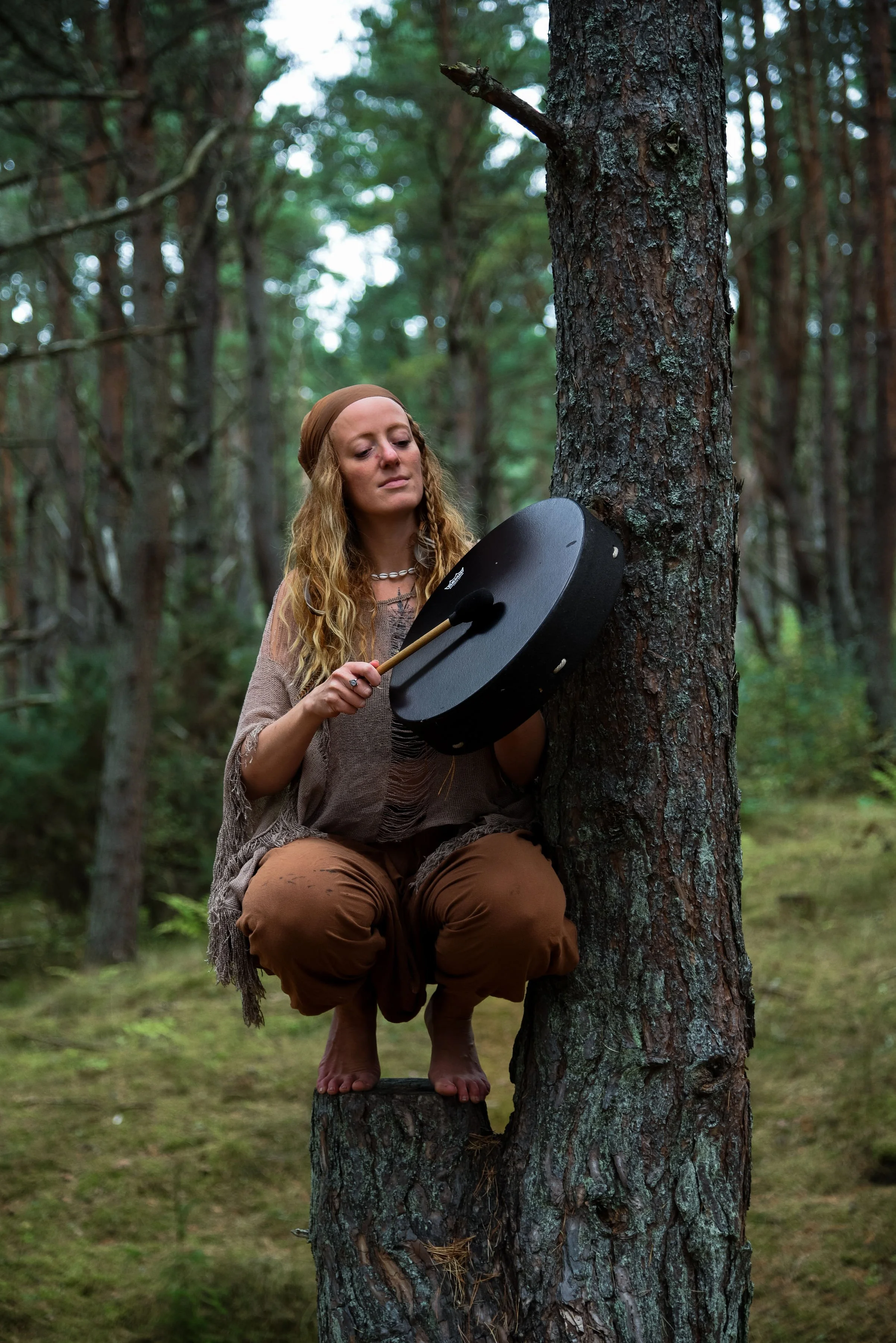 A woman with long blonde hair, wearing a headband, brown pants, and a loose, brown top, is squatting on a tree stump in a forest. She is holding a black gong and a mallet, preparing to strike the gong.